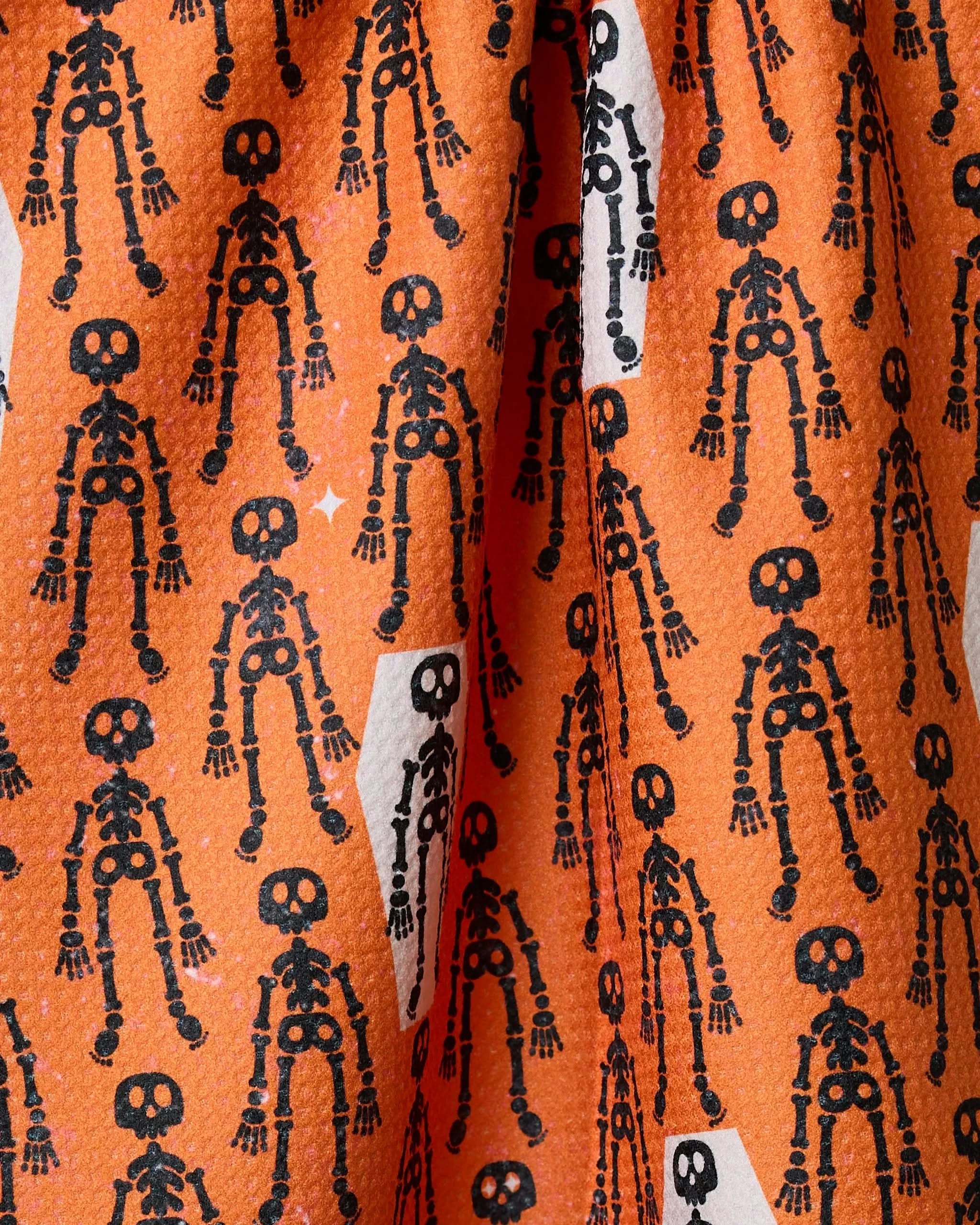 Close-up of Geometrys Resting Skeletons Orange tea towel, made from recycled materials, featuring a playful pattern of black skeletons and white stars on an orange background.