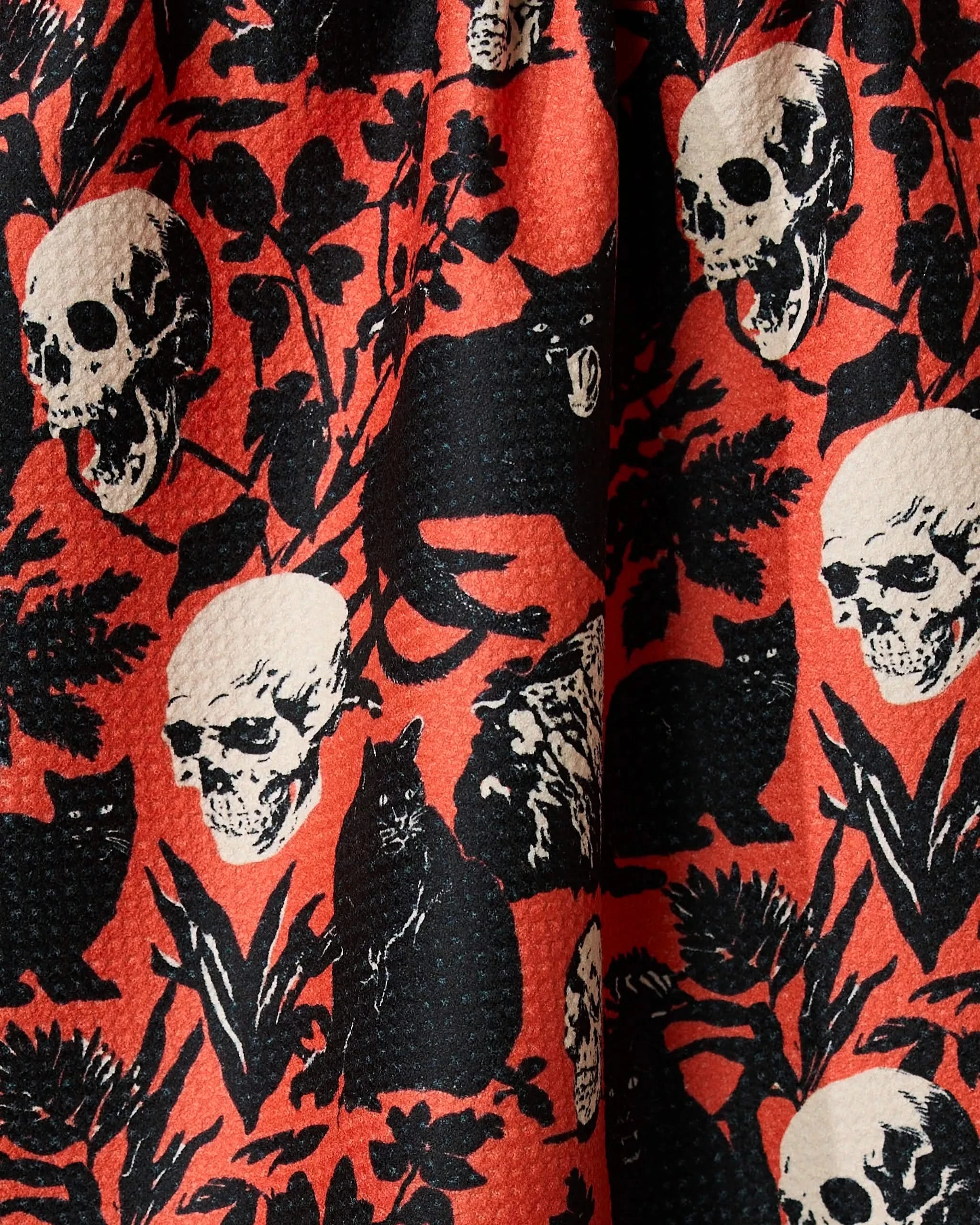 The Geometry It Comes At Night tea towel features a bold, spooky textured orange design with black cats, white skulls, and black plants, made from recycled materials.