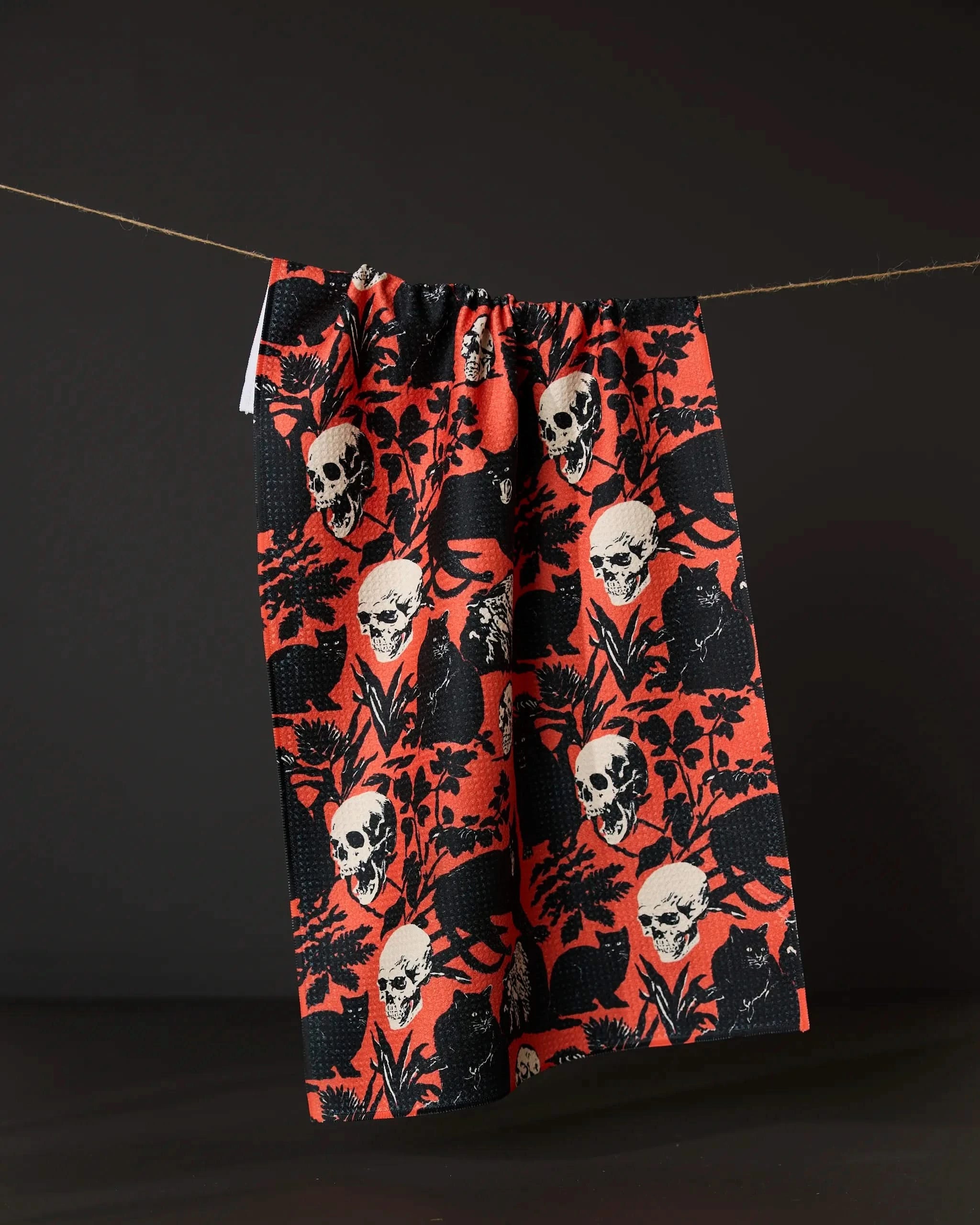 The Geometry It Comes At Night tea towel, made from recycled materials and featuring red and black skull, cat, and leaf patterns, hangs from a string against a dark background.