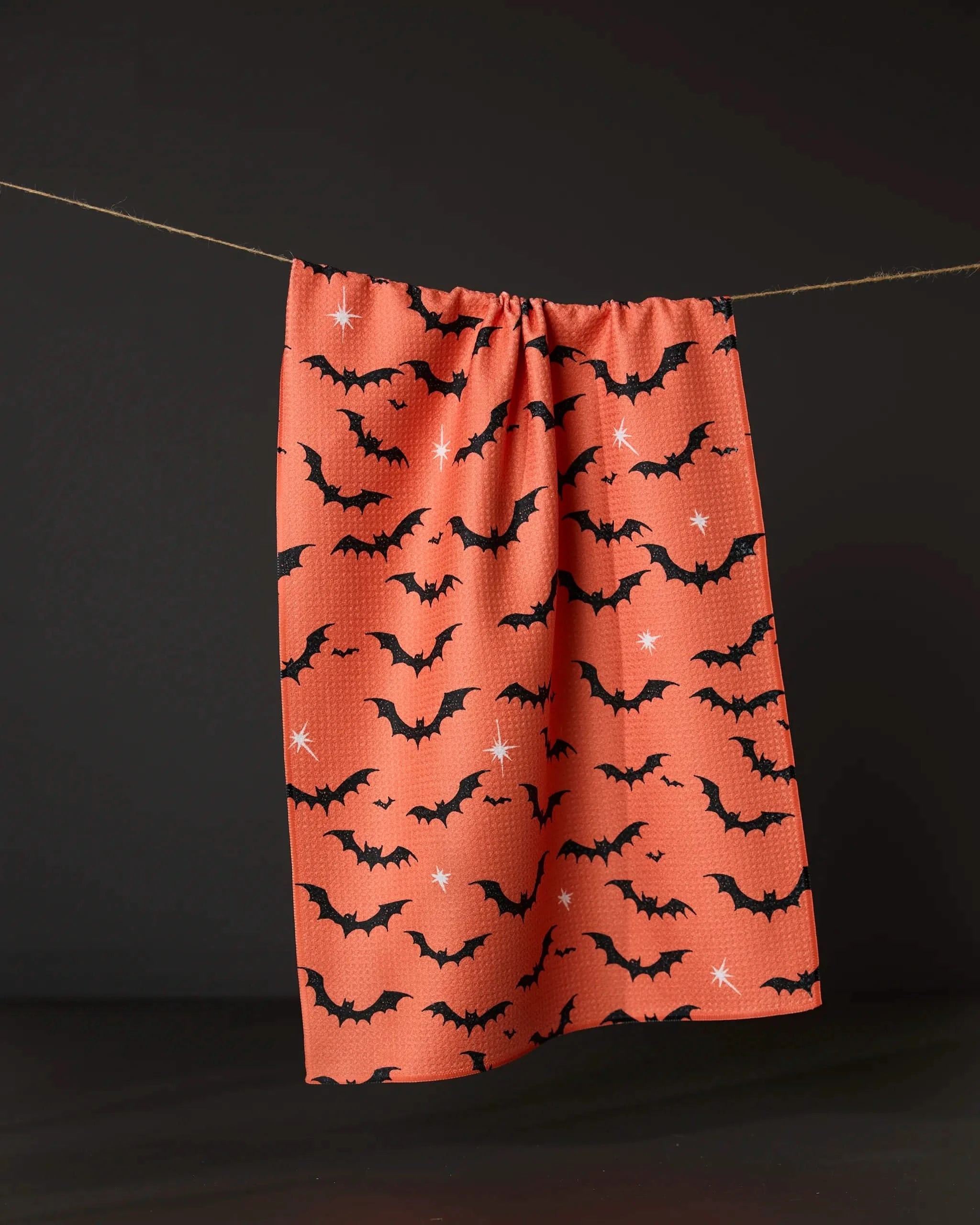 The Geometry Midnight Friends Orange tea towel, made from recycled materials, features black bats and white stars on orange fabric and is displayed against a dark background.