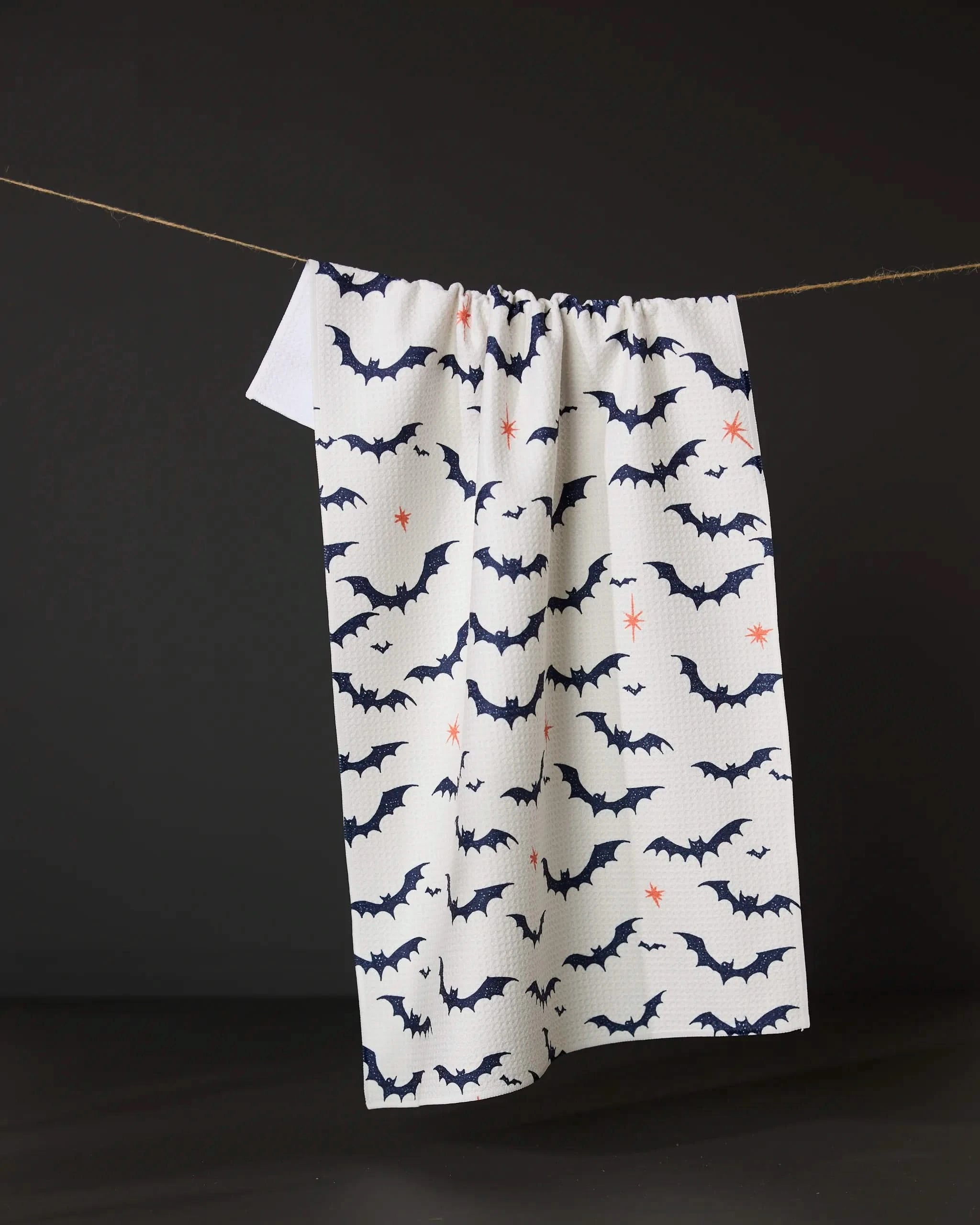 The Geometry Midnight Friends White dish towel, featuring dark blue bats and small orange stars, hangs on a string against a black background.