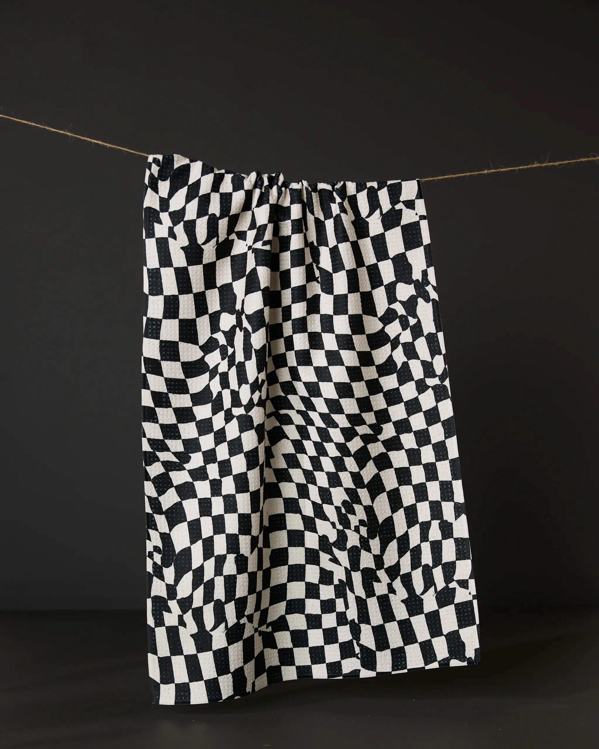 The Glitchy Check tea towel by Geometry, featuring a black and white wavy checkered design, hangs against a dark background. Made from recycled materials.