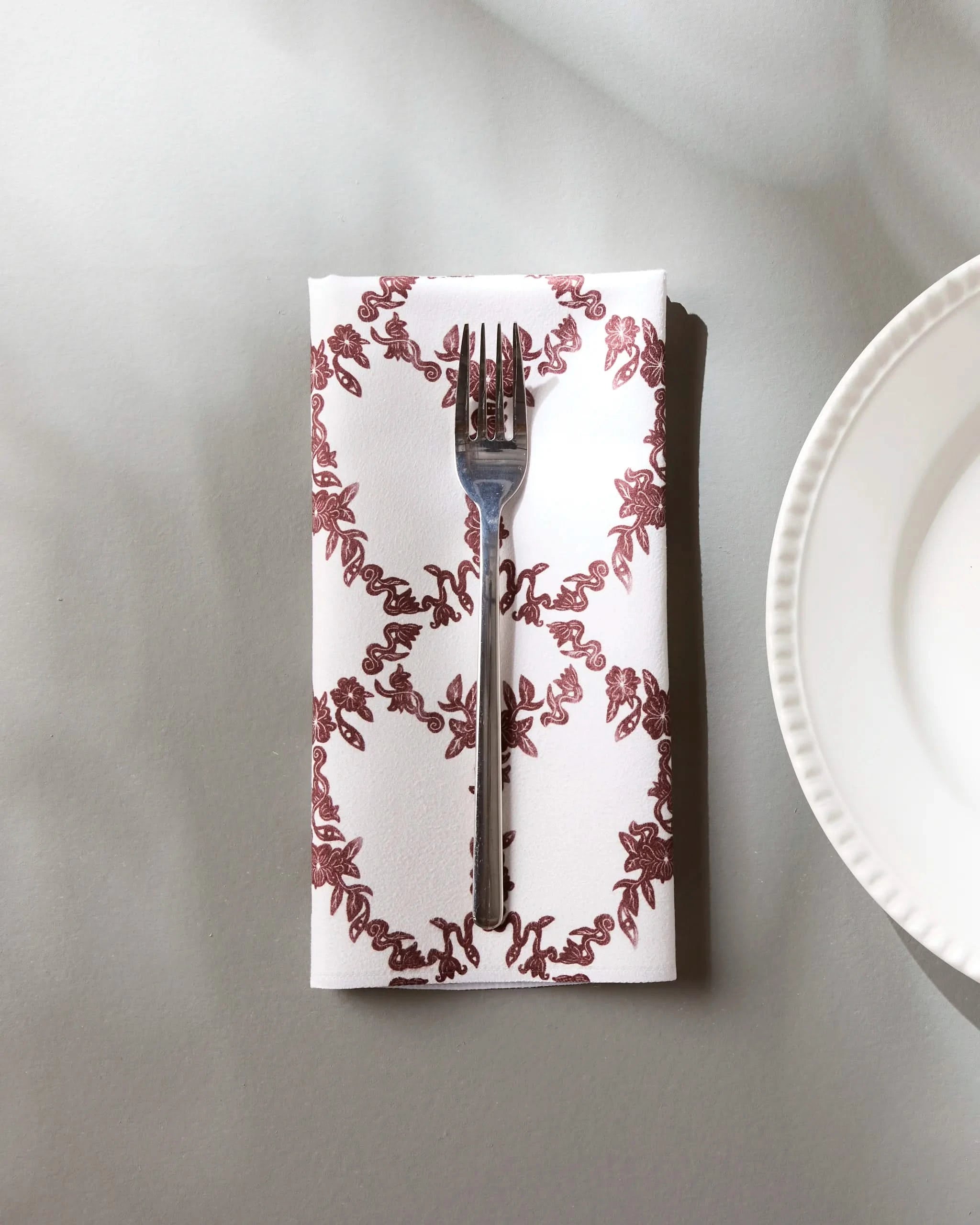 A silver fork sits on the Geometry Eye on You Floral eco-friendly napkin, which features a decorative maroon border, placed on a light gray table beside a white ridged dinner plate.