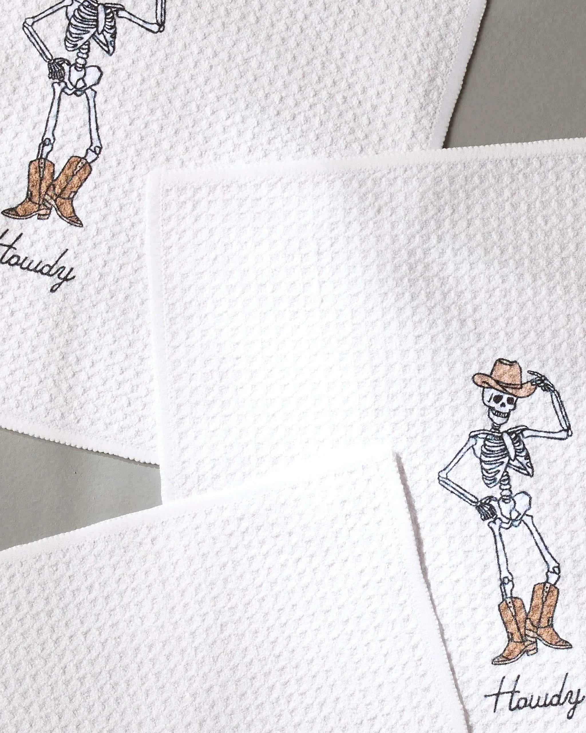 Set of three white textured Geometry Howdy Cassondra dishtowels, each featuring a unique embroidered skeleton in cowboy hats and boots with Howdy stitched at the bottom. Super-absorbent and stylish for your kitchen.