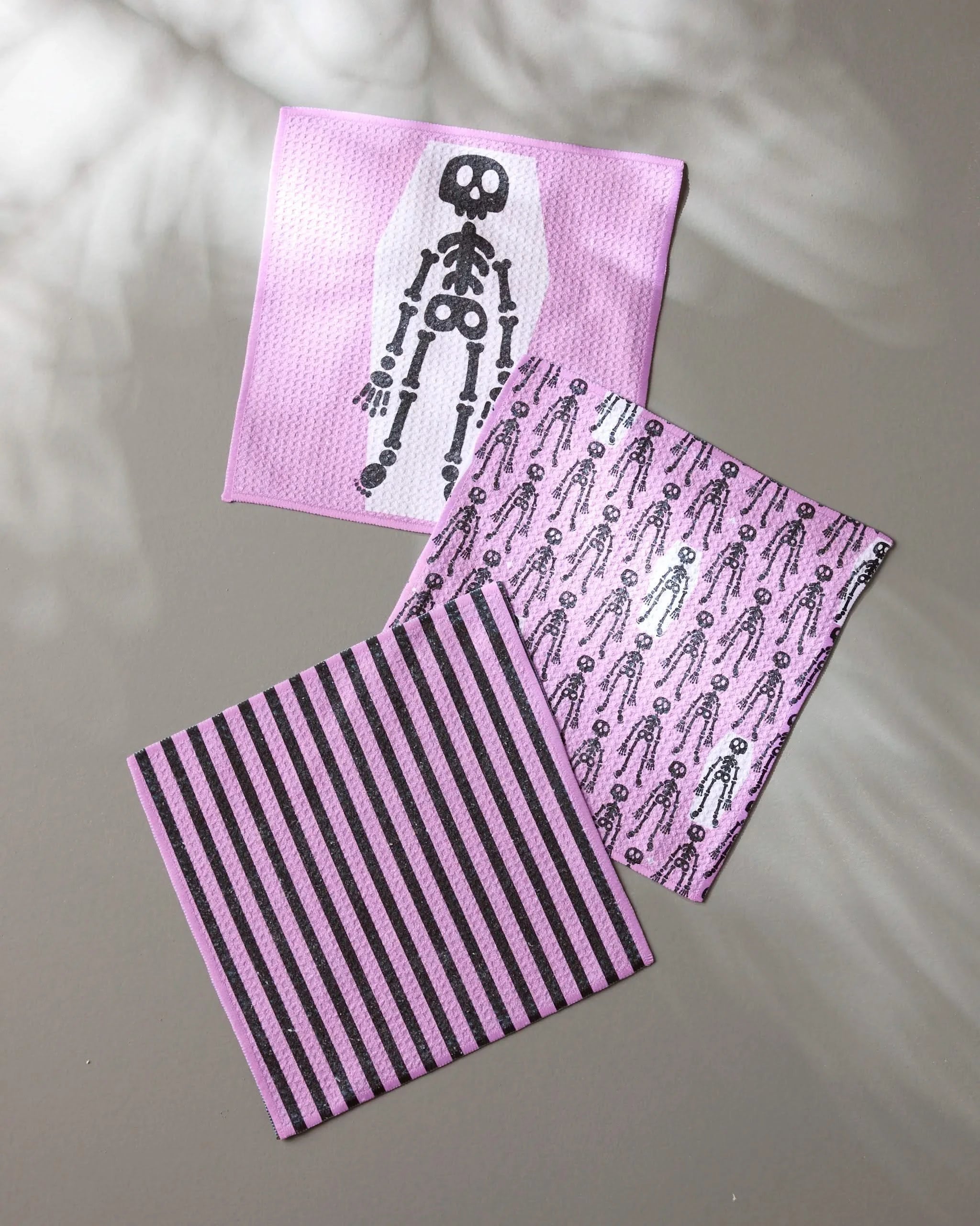 The Geometry “Bright Bones” set features three recycled dishcloths—one with a skeleton graphic, one with a skeleton pattern, and one striped—all in lavender, their details highlighted as light and shadow play across the gray surface.