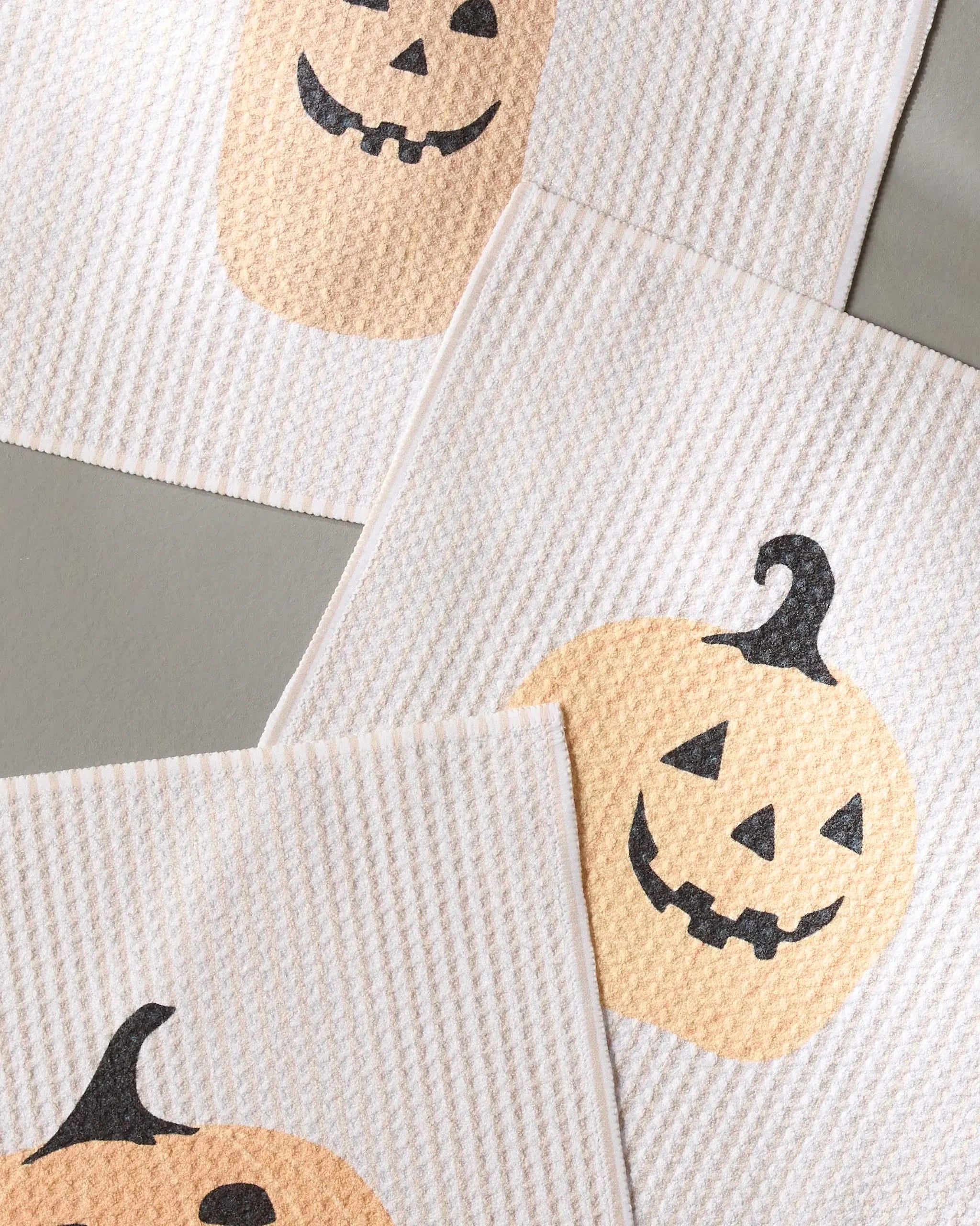 The Gourdgeous dishcloth set by Geometry includes three white textured cloths made from recycled materials, each with smiling orange jack-o-lantern faces and black stems—perfect for adding a festive touch to your kitchen.