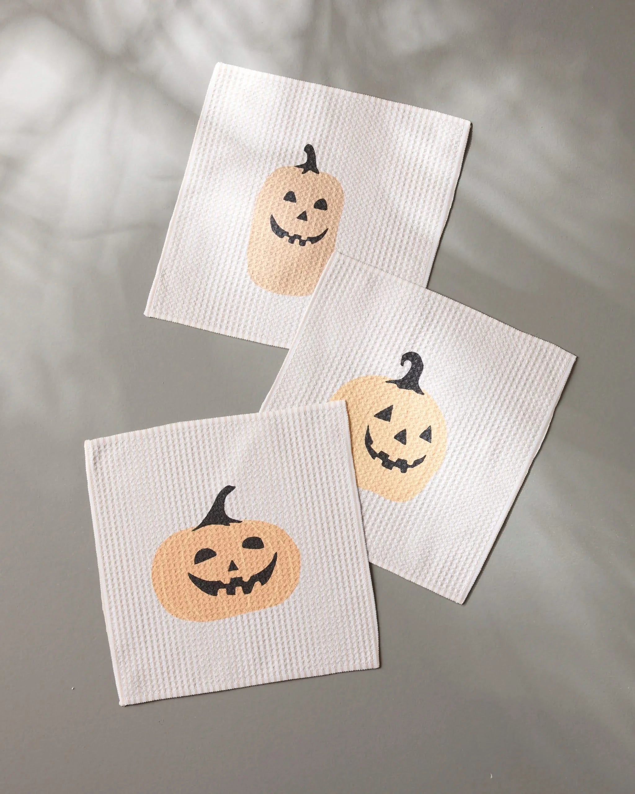 Three Gourdgeous white textured napkins by Geometry, made from recycled materials and featuring smiling jack-o-lantern faces, are arranged on a gray surface with soft shadows.