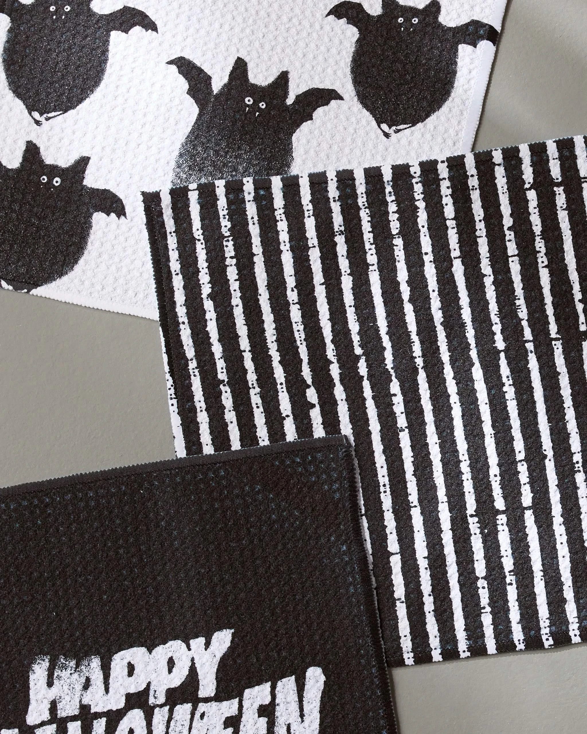 The Geometry Macabre Halloween-themed set includes three absorbent dishcloths—one with black bats, one with black and white stripes, and one with Happy Halloween in bold white letters—all made from recycled materials.