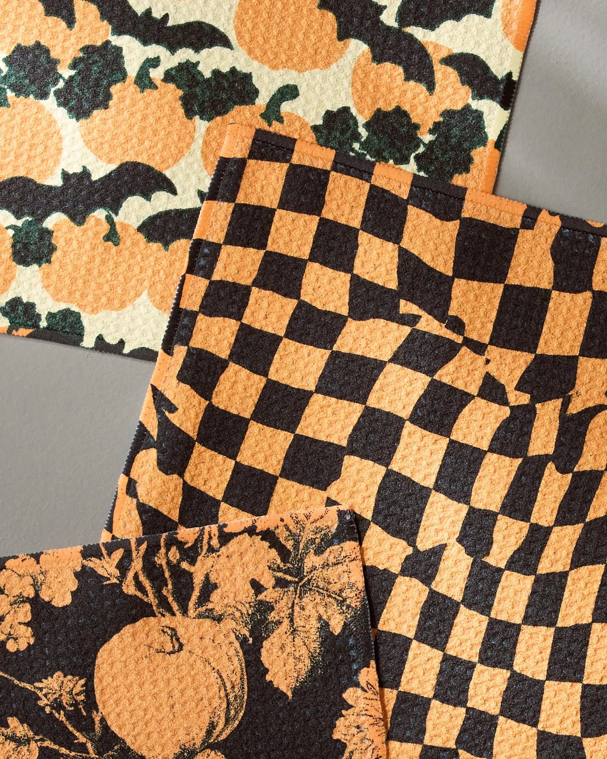 The Geometry Night Ride Halloween dishcloth set features three fast-drying towels with festive pumpkins, bats, a wavy checkerboard, and pumpkin leaf designs, arranged overlapping on a gray surface.
