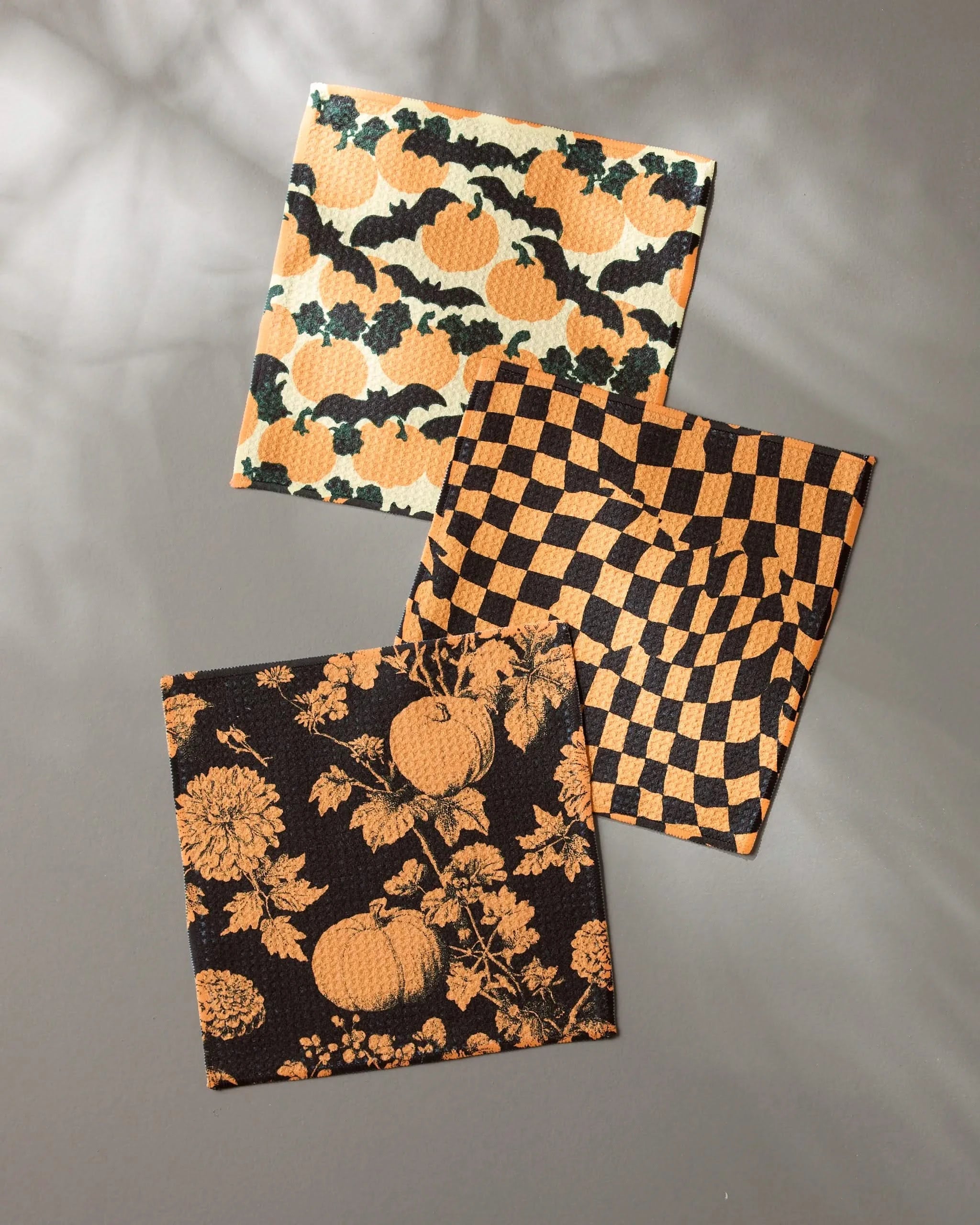 Three square autumn-themed dishcloths from Geometrys Night Ride set: made from recycled materials and featuring orange pumpkins with black leaves, an orange and black check pattern, and pumpkins with flowers on black.