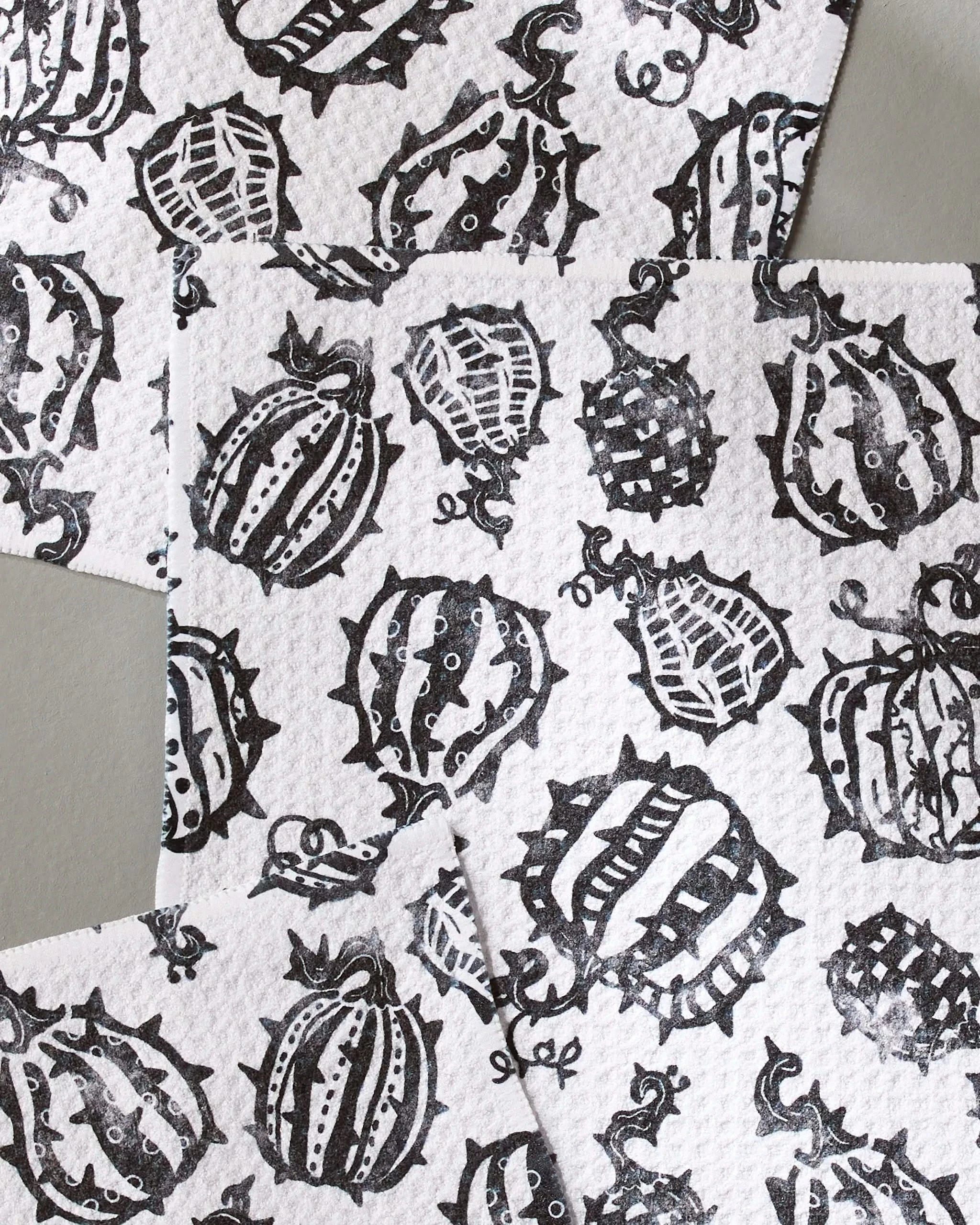 Geometry’s Punk Pumpkins includes three white textured dishcloths made from recycled materials, each with black hand-drawn gourds and pumpkins overlapping on a light gray background.