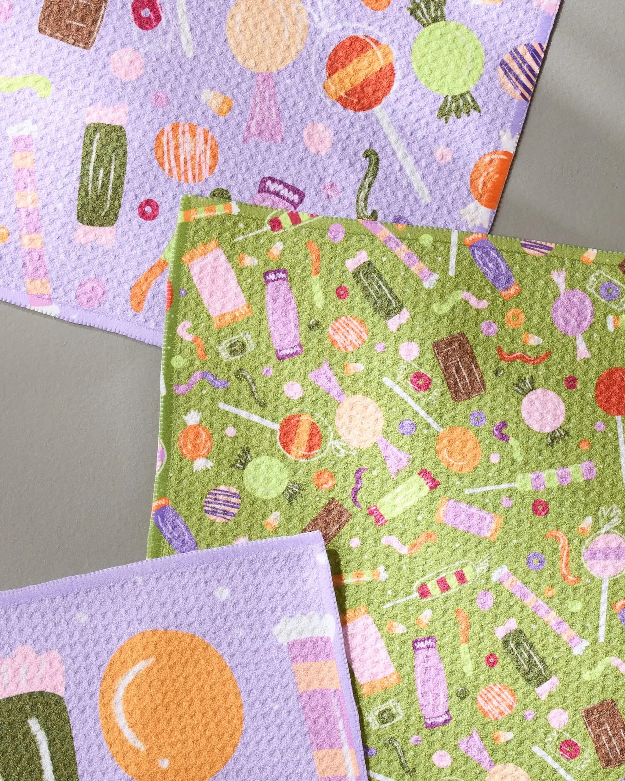 Geometrys Sweet Treasures set features three ultra-absorbent dishcloths with vibrant candy patterns on purple, green, and lilac backgrounds, arranged on a gray surface.