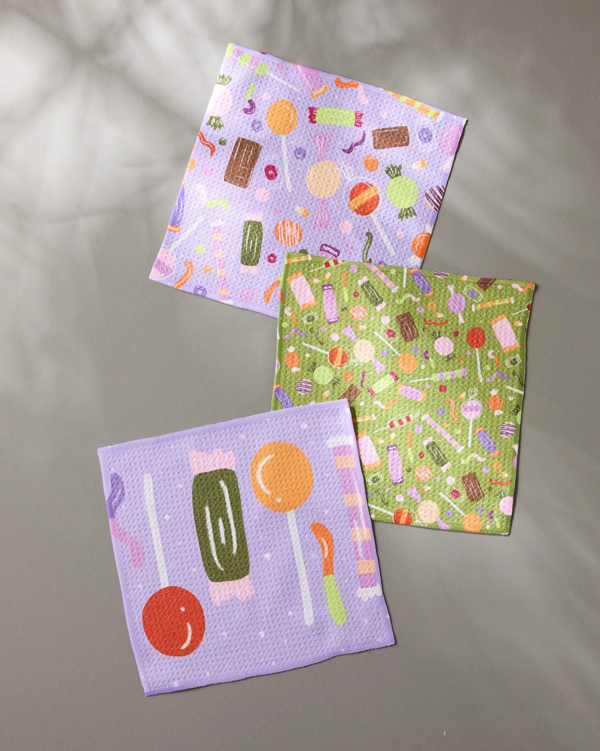 Three Sweet Treasures dishcloths from Geometry—one green, one purple, and one lavender with candy-inspired designs—are arranged on a gray surface bathed in dappled sunlight and shadow.
