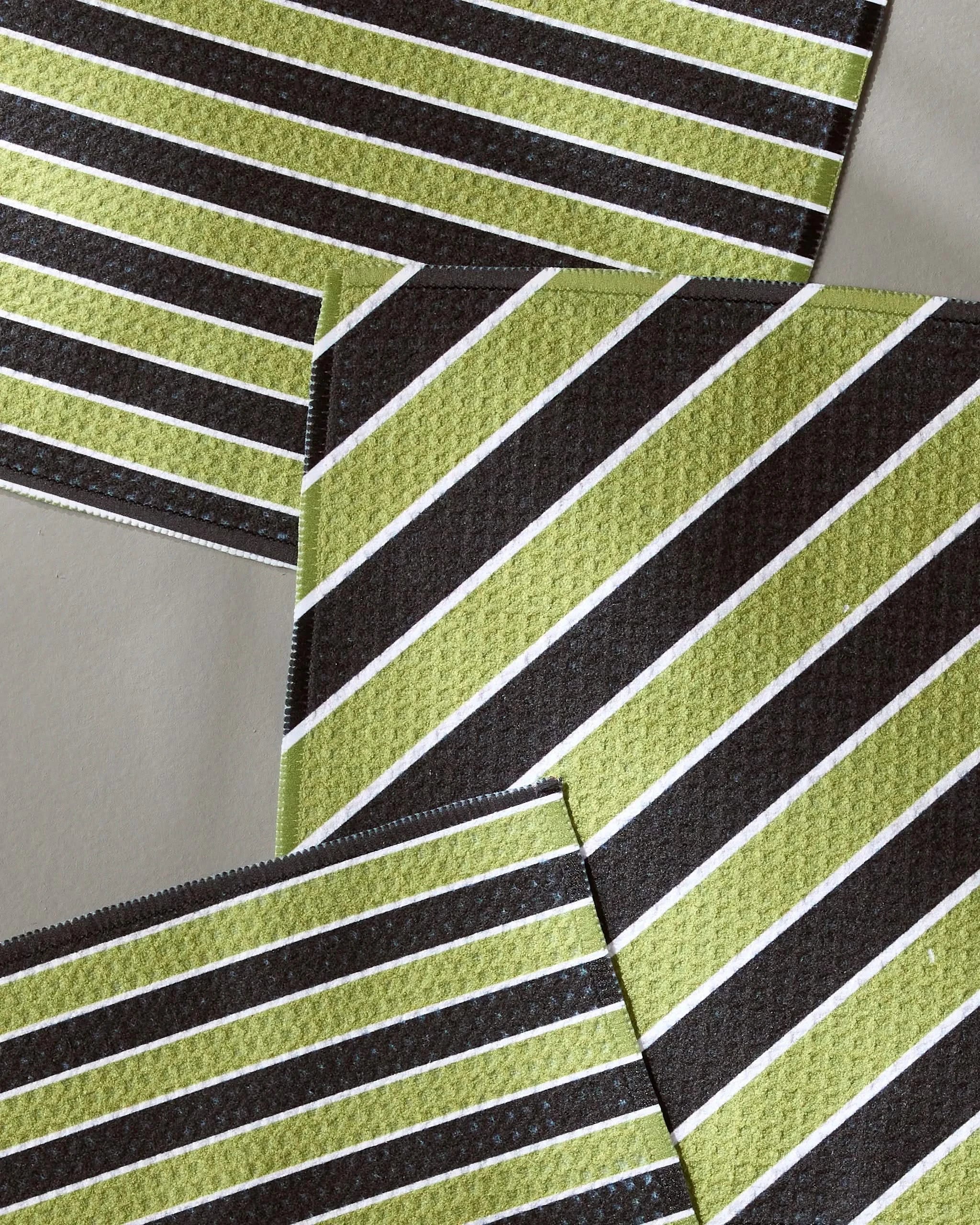 Four overlapping Tricky Lines fabric swatches by Geometry, featuring green, black, and white diagonal stripes made from recycled materials, are arranged on a gray background for a modern geometric look.