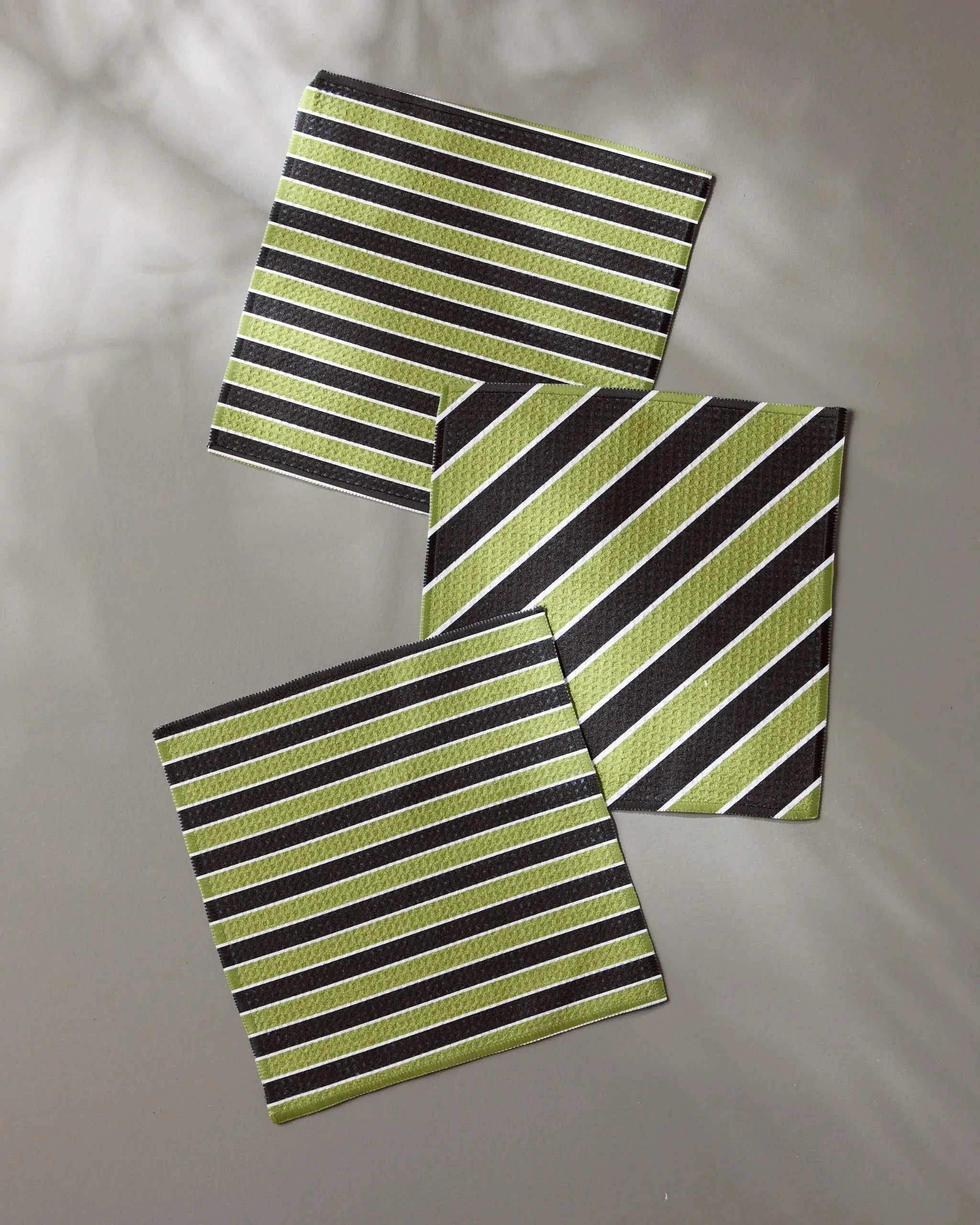 Geometry’s Tricky Lines: a set of three square coasters crafted from recycled materials, each with green, white, and brown stripes in diagonal and horizontal designs, displayed on a gray surface.