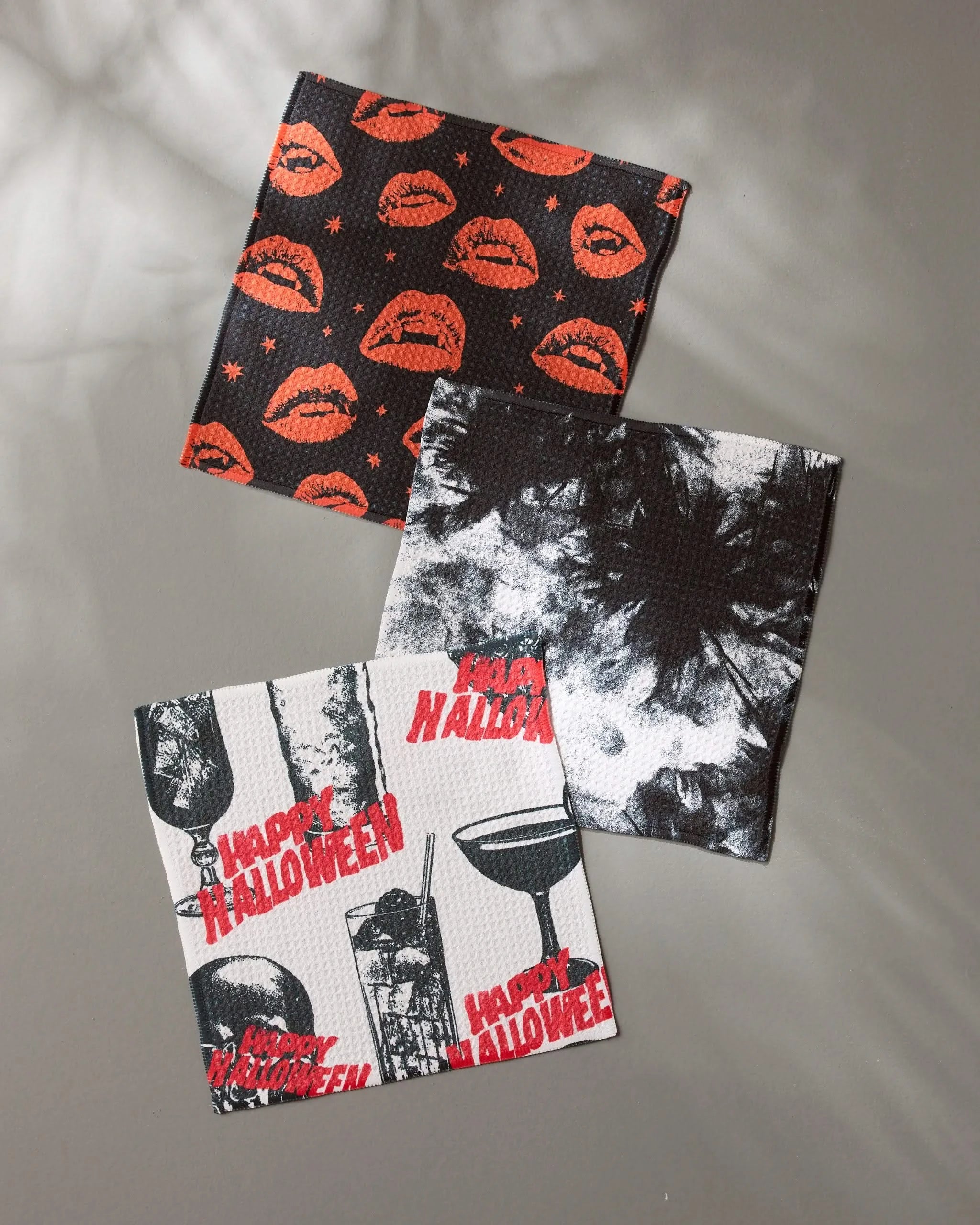 The Geometry Witchin Hour set includes three square, super-absorbent dishcloths with Halloween-themed prints: orange lips on black, black and white tie-dye, and “Happy Halloween” with spooky red and black designs on white.