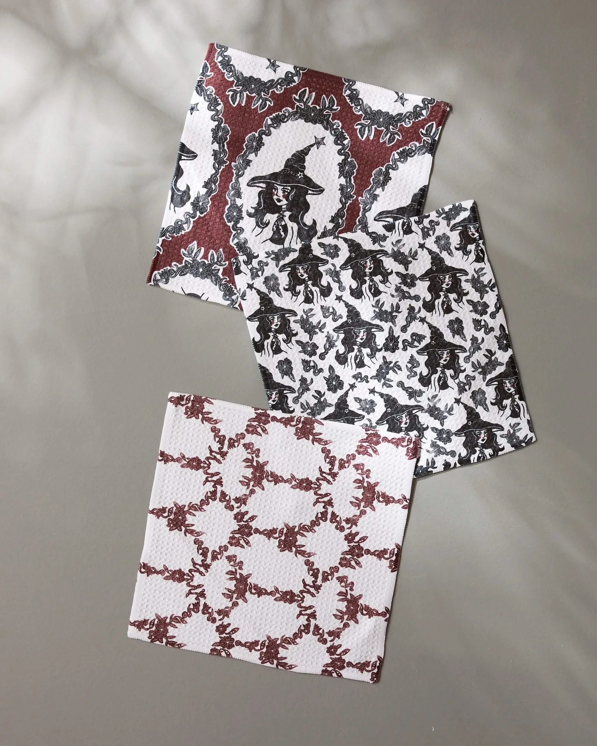 Three square Witchy Blooms napkins by Geometry, made from recycled materials, display intricate red and black witch and floral patterns. They overlap on a neutral surface with soft light and subtle leaf shadows.