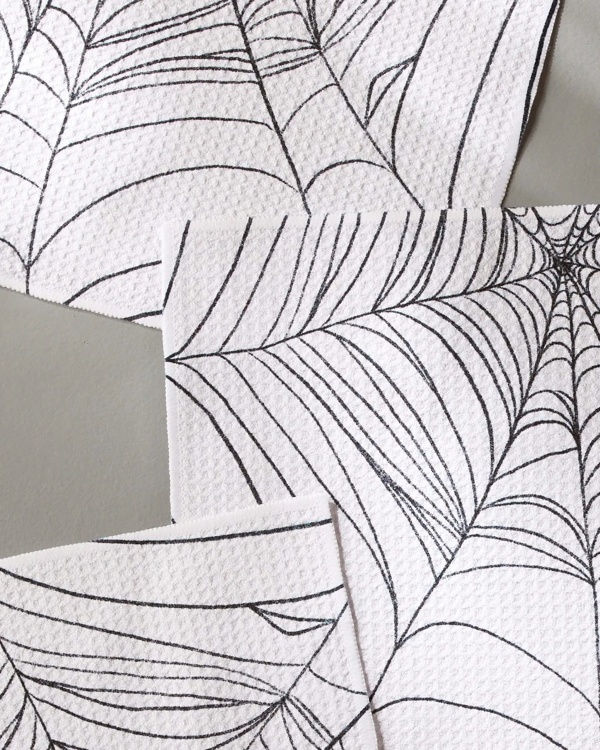 Three Webbed Out napkins from Geometry overlap on a light gray surface. Each white napkin features thin, stylized black spiderweb designs and is crafted from recycled materials.