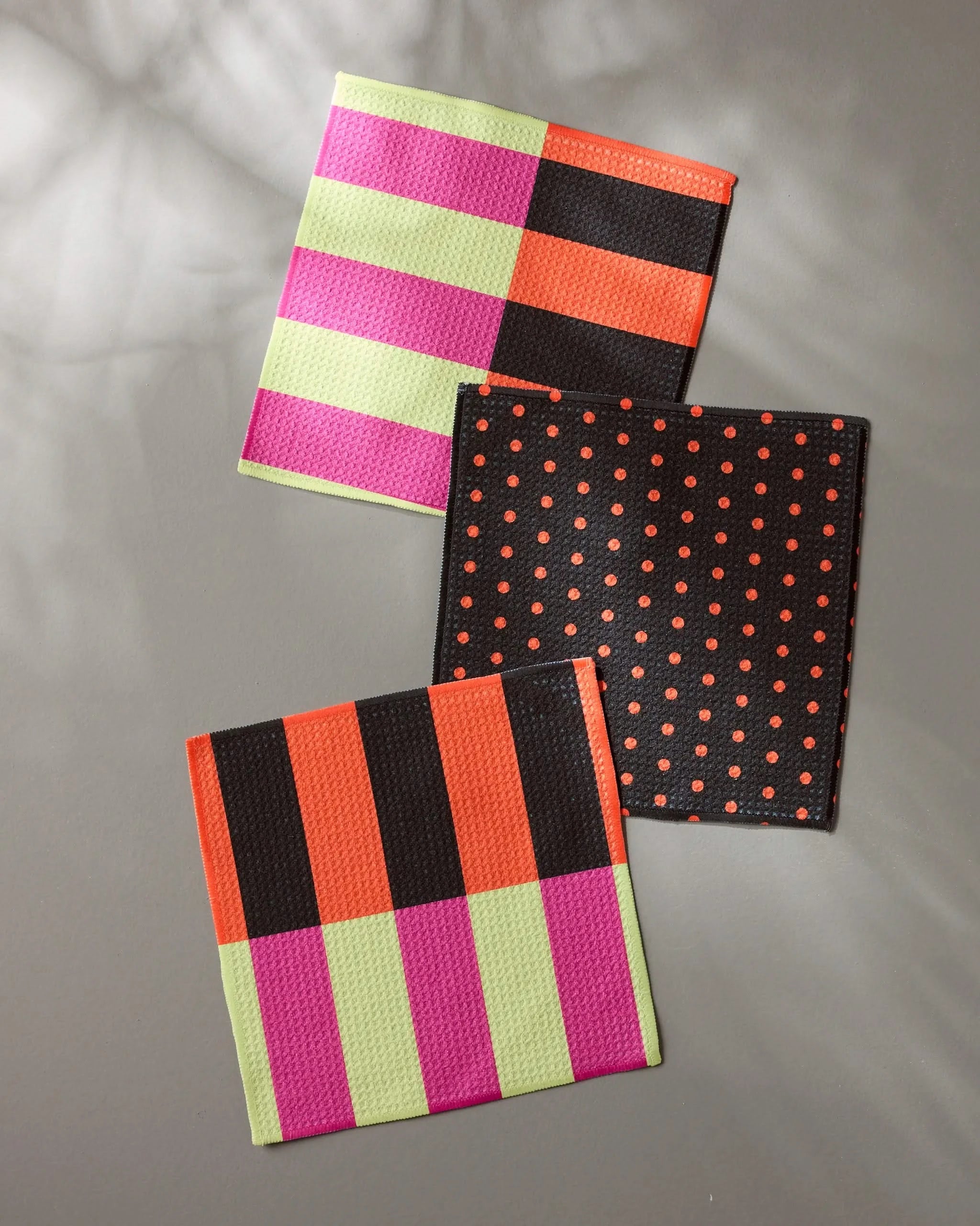 Three colorful Geometry Spooky Hour Stripe coasters rest on a gray surface—perfect with a super-absorbent dish towel set. Designs feature pink, green, orange, and brown stripes; orange polka dots; and playful blocks under soft lighting.