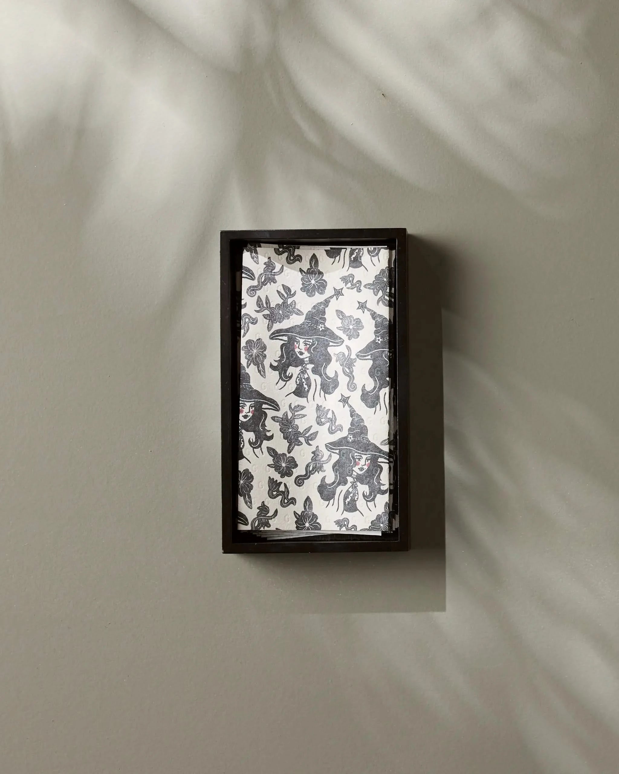 Geometry’s Floral Witches features a black-framed rectangular box with a black and white abstract interior, mounted on a light gray wall with leafy shadows, evoking the subtle designs of eco-friendly kitchen towels.