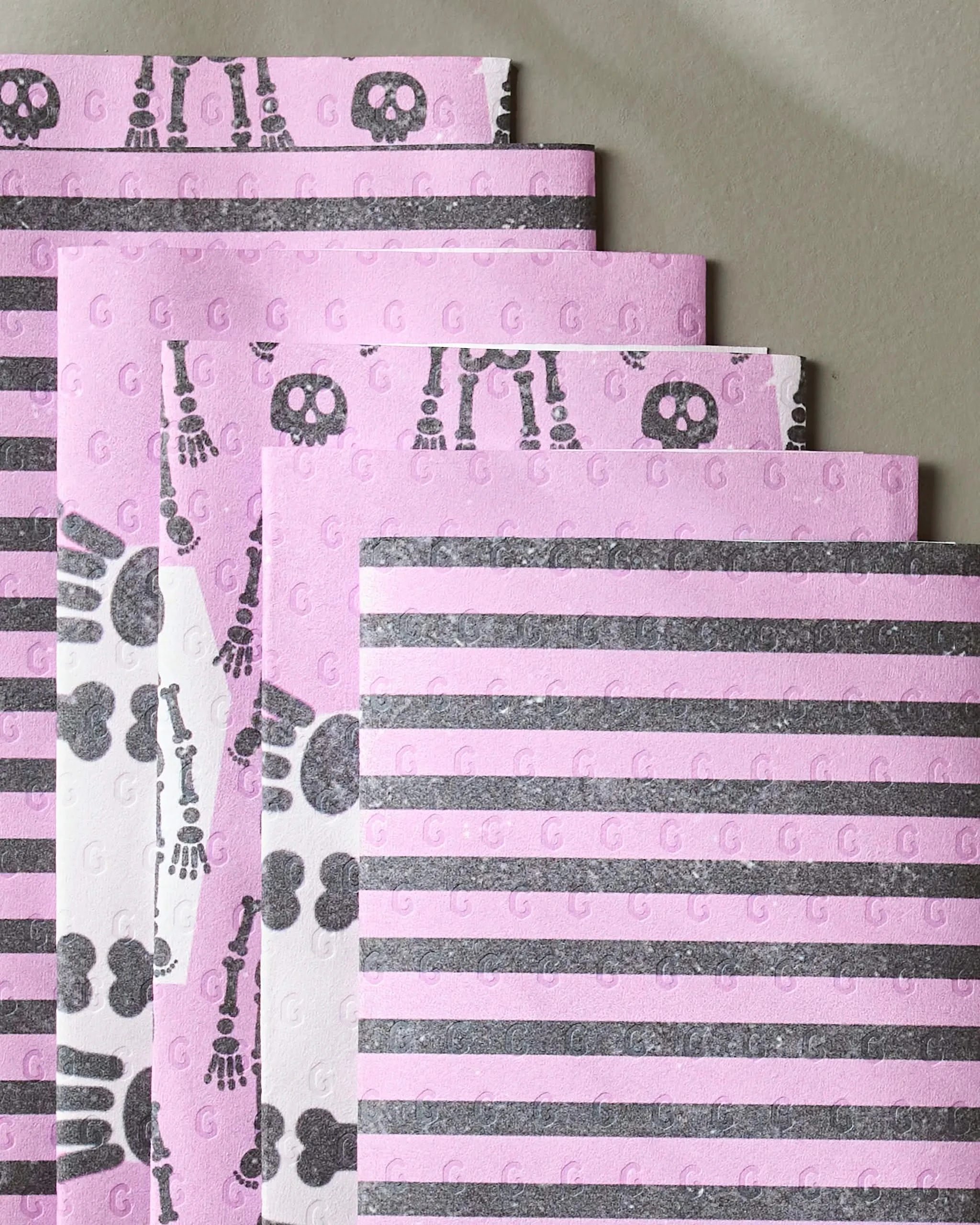 Geometry’s Bright Bones features four eco-friendly paper towel sheets in pink and gray, each with horizontal stripes, skeleton bones, or skull designs. The sheets are slightly overlapped on a neutral background.