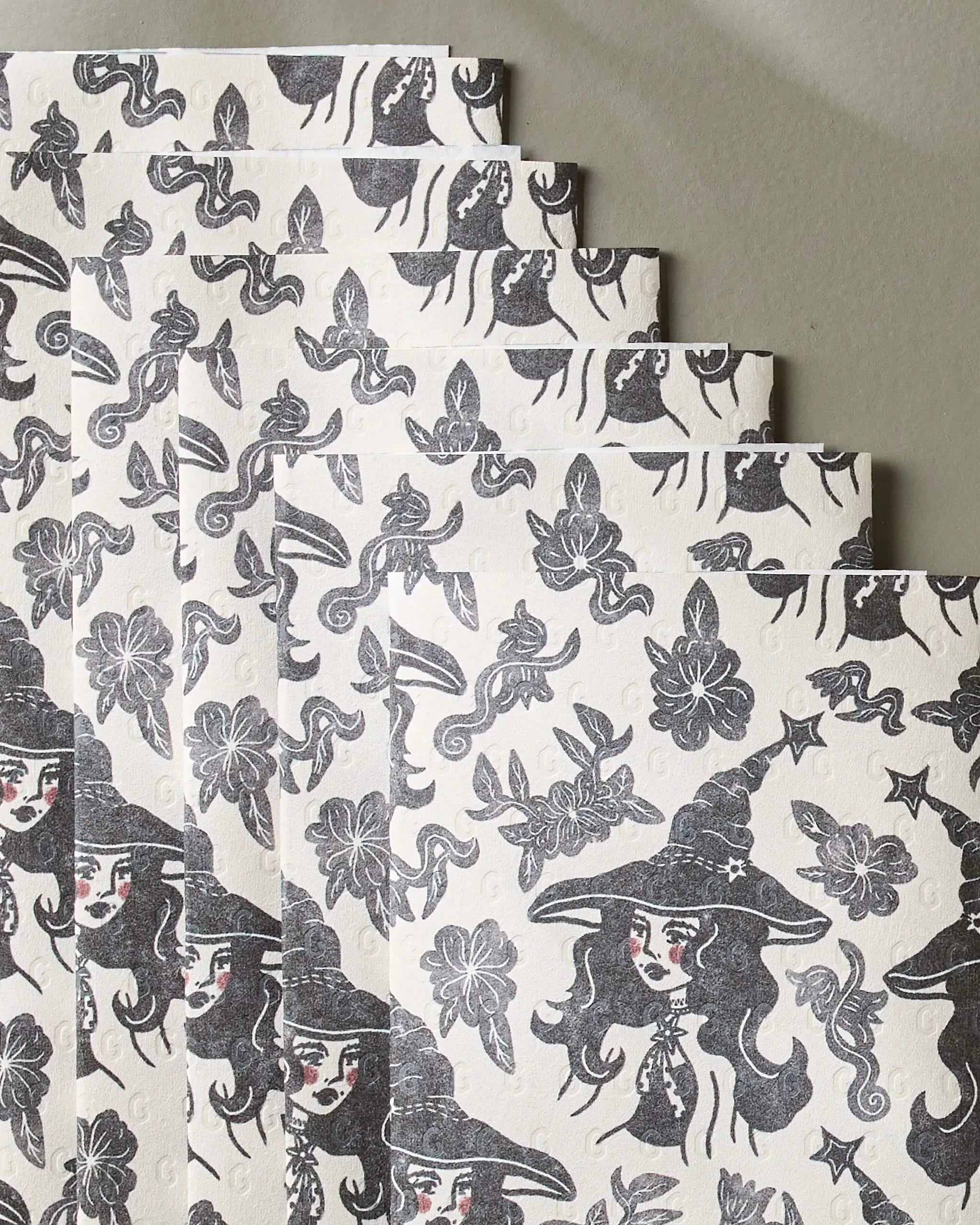 Several sheets of Geometry’s Floral Witches eco-friendly kitchen towels, showcasing black and white designs with witch faces in hats, snakes, bats, flowers, and crescent moons, are arranged in a staggered overlap.