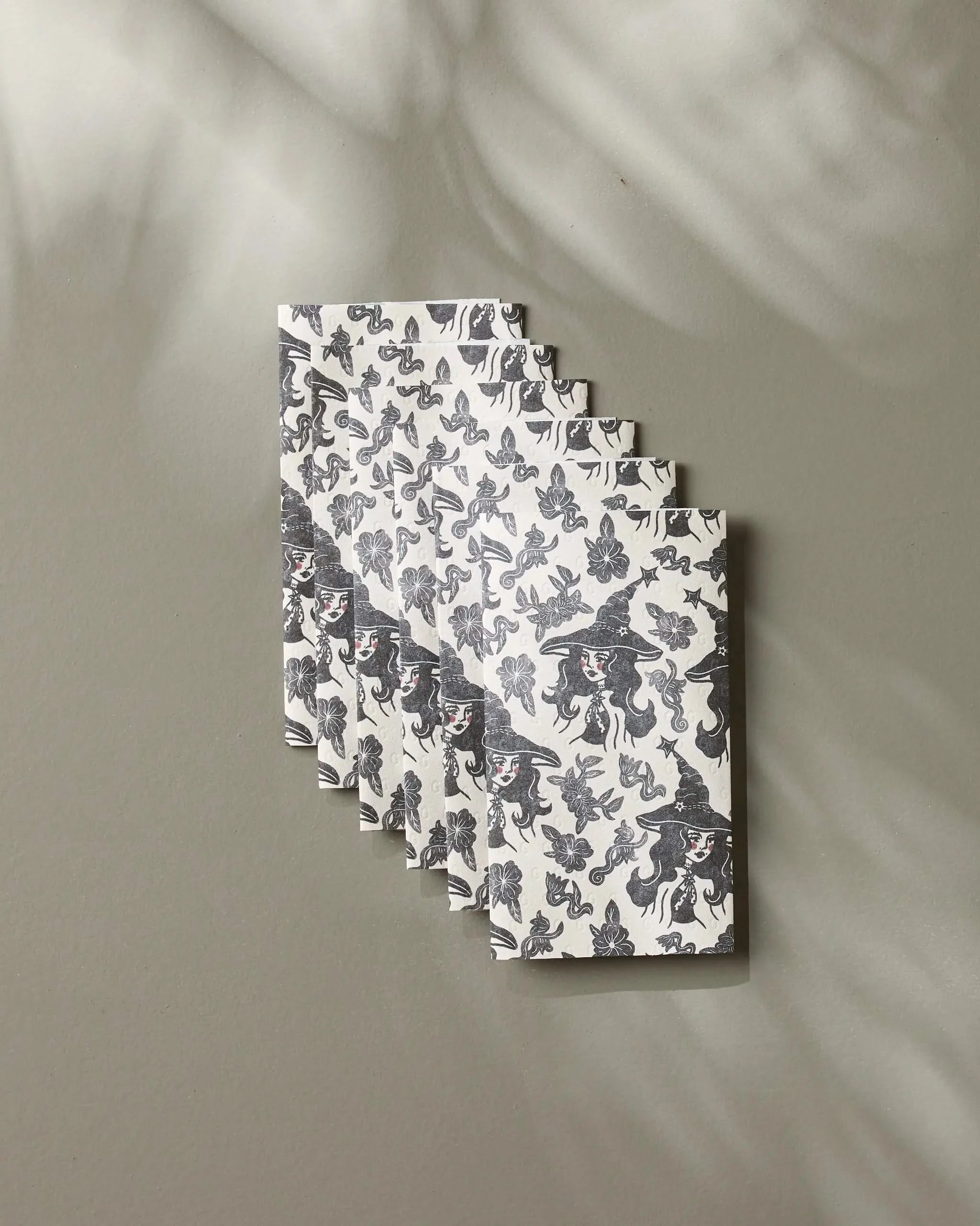 Five “Floral Witches” cards by Geometry, featuring a black and white witch and butterfly pattern, are fanned on a gray surface with soft shadows, their enchanting design recalling eco-friendly kitchen towels.