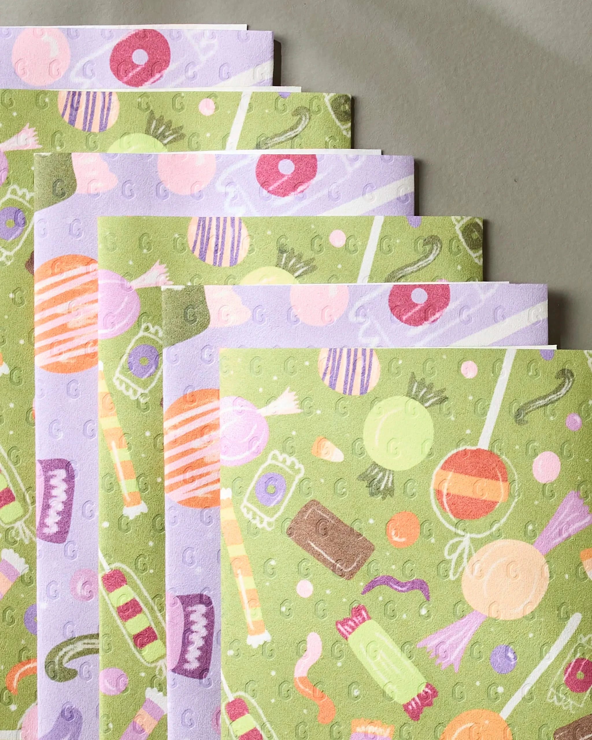 Five eco-friendly Geometry Sweet Treasures paper towels, featuring colorful assorted candies, lollipops, licorice, and chocolates on a green and lavender background, are fanned out on a neutral surface.