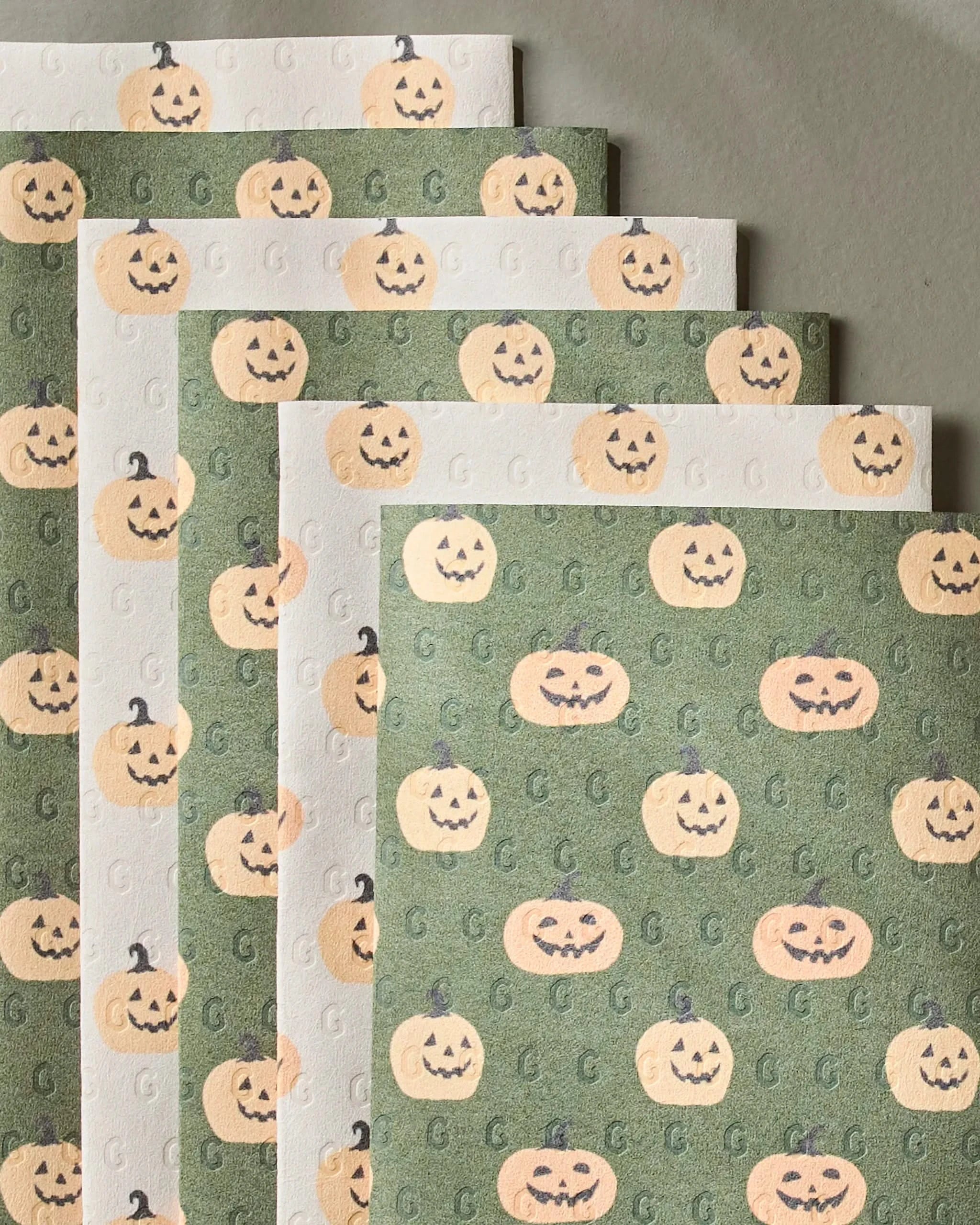 Spice and Smiles by Geometry features sheets with jack-o’-lantern pumpkin patterns on green and off-white backgrounds, fanned out and ideal as a recycled paper towel alternative for festive occasions.