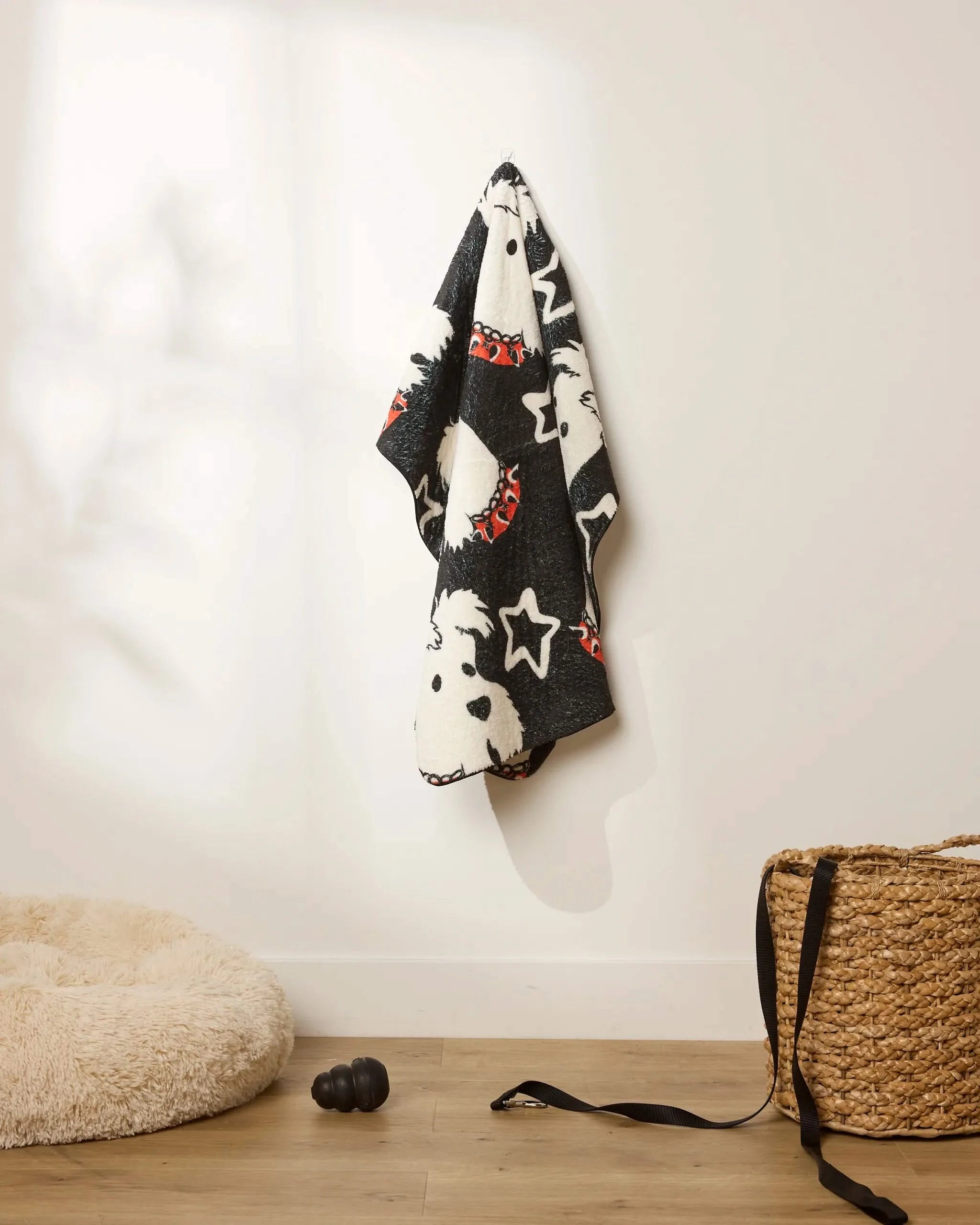 The Geometry Happy Howleen black and white plush dog towel, featuring cartoon dog and star designs, hangs on a white wall above a wooden floor beside a round pet bed, woven basket, and black dog toy.