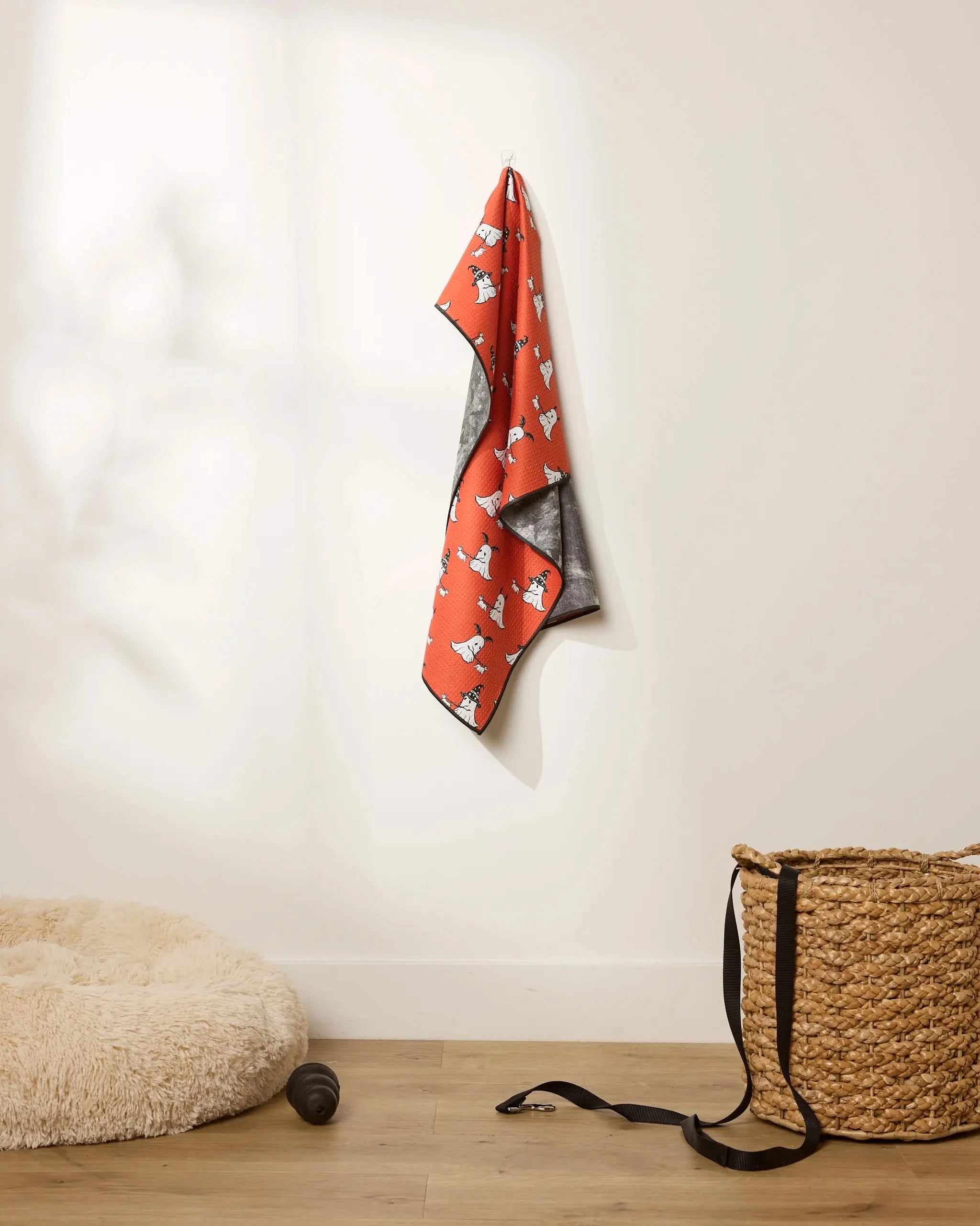 The Geometry Spooky Ghost Walk Orange, a highly absorbent dog towel with charming prints, hangs on a white wall above a wooden floor. Nearby are a fluffy dog bed, a black toy, and a woven basket. Soft natural light creates gentle shadows.