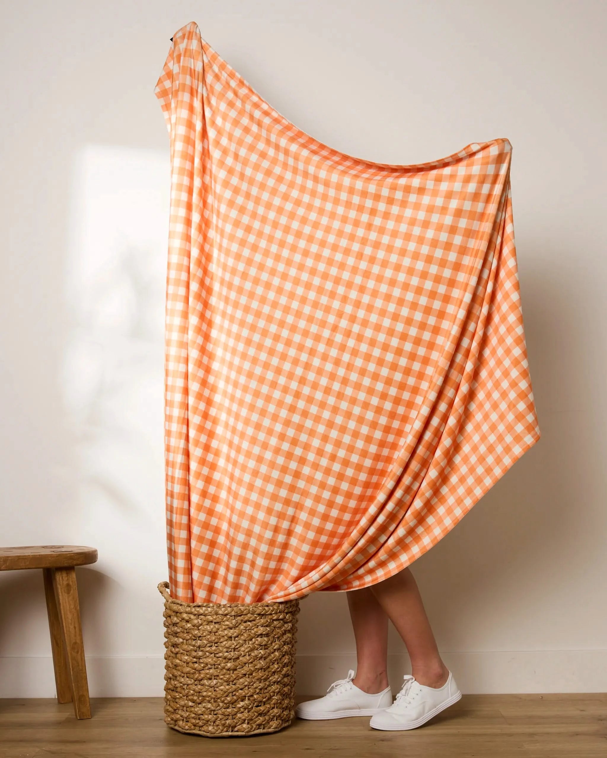 A person stands in a woven basket, mostly hidden behind the Geometry Boohoo Orange gingham throw draped over their head. Only their legs and white sneakers are visible, with a wooden stool nearby against a plain wall.
