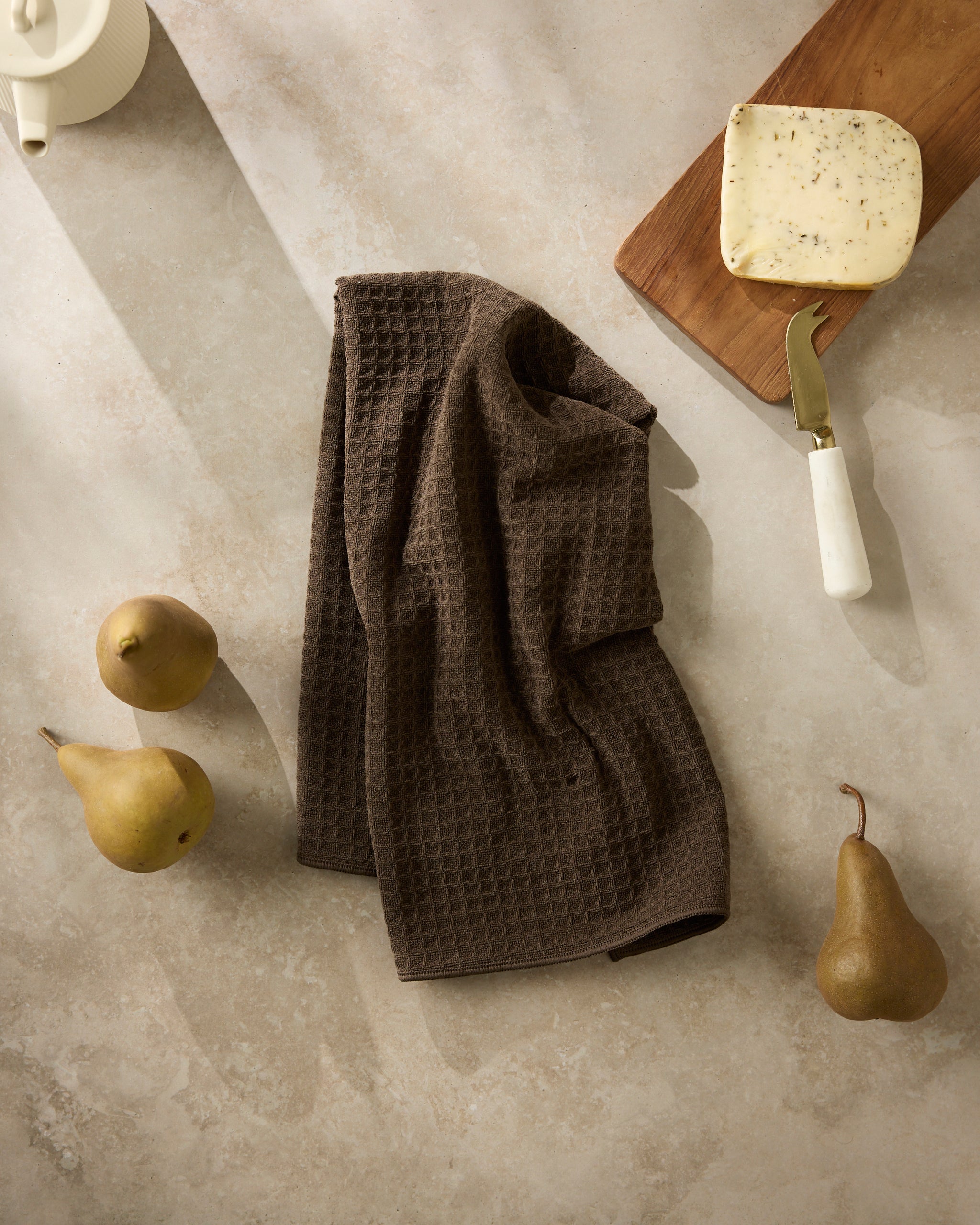 A Geometry “Espresso” brown textured kitchen towel is spread on a light stone surface, with three pears, a herb cheese block on a wooden board, and a white-handled cheese knife. Sunlight casts soft shadows across the scene.