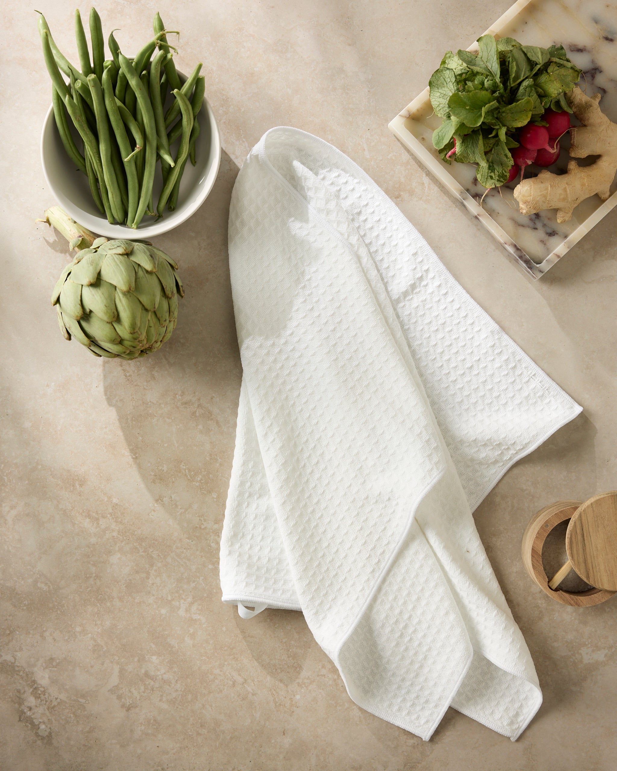 A white textured Geometry Salt kitchen towel lies on a countertop next to a bowl of green beans, an artichoke, a marble tray with radishes, greens, ginger, and a small wooden object.