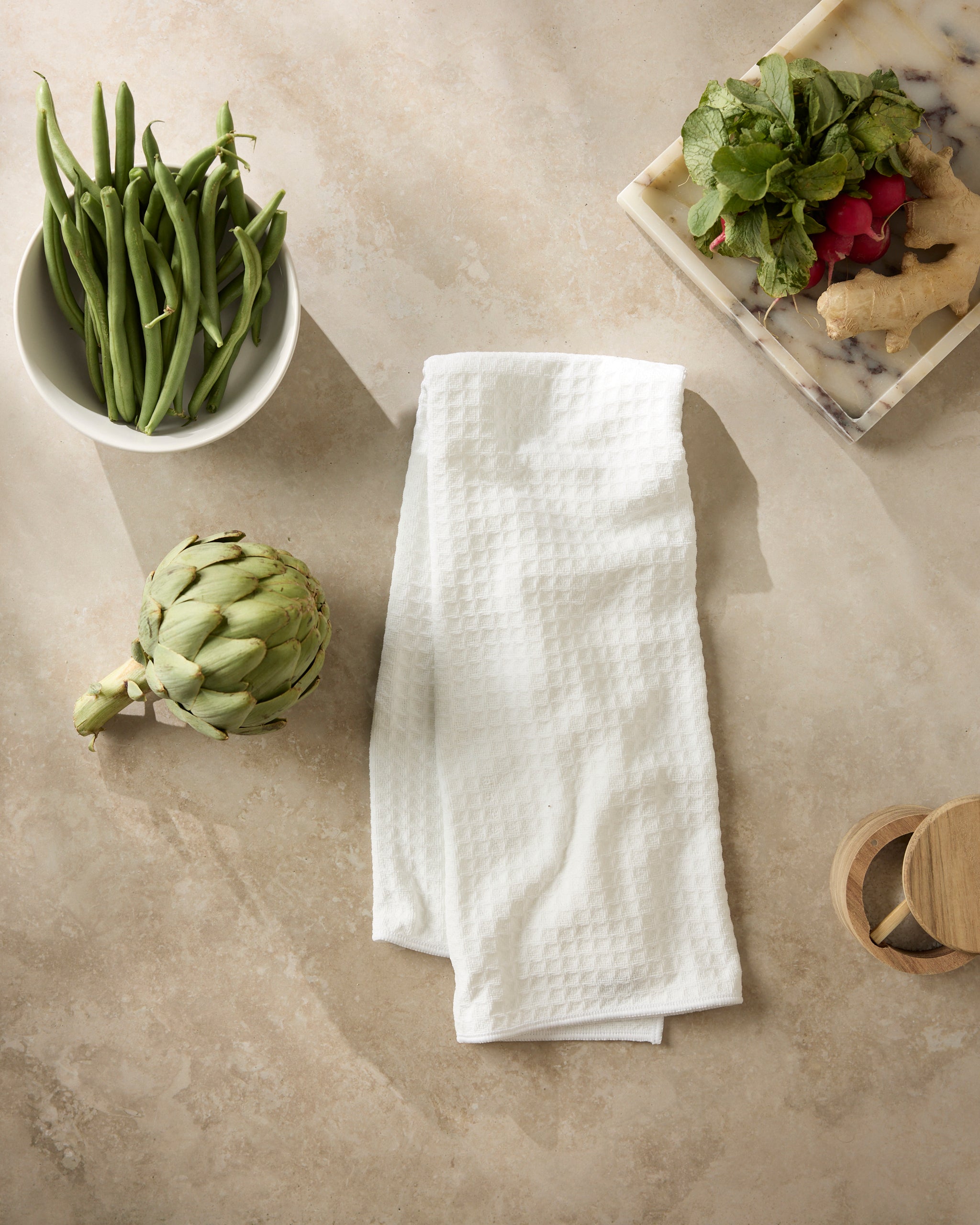 A Geometry Salt white textured kitchen towel is spread on a light countertop, with a bowl of green beans, an artichoke, a marble tray of radishes, ginger, greens, and two small wooden containers surrounding it.