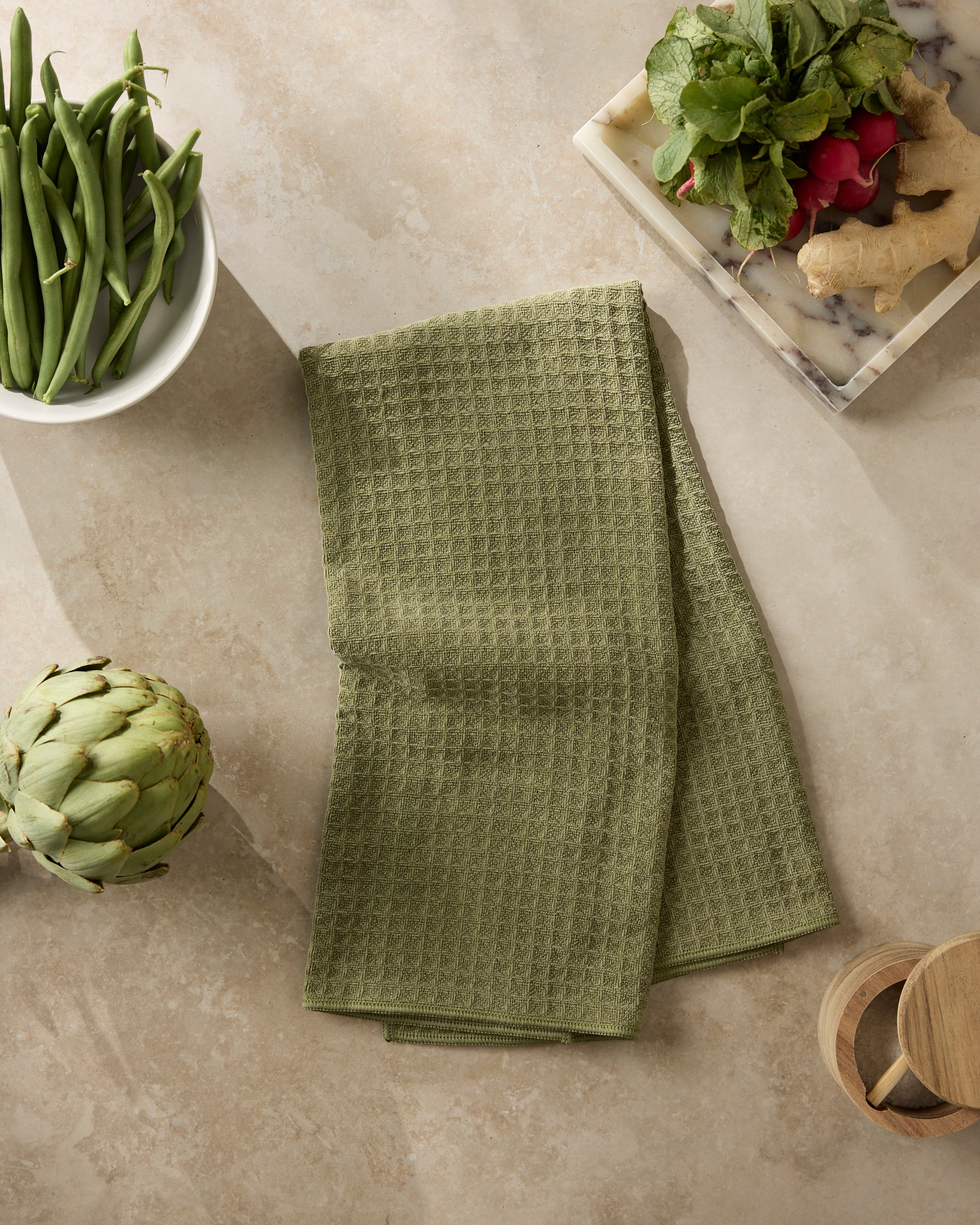 A folded Geometry Olive kitchen towel rests on a beige countertop, surrounded by a bowl of green beans, an artichoke, a marble board with ginger and radishes, and two wooden containers.