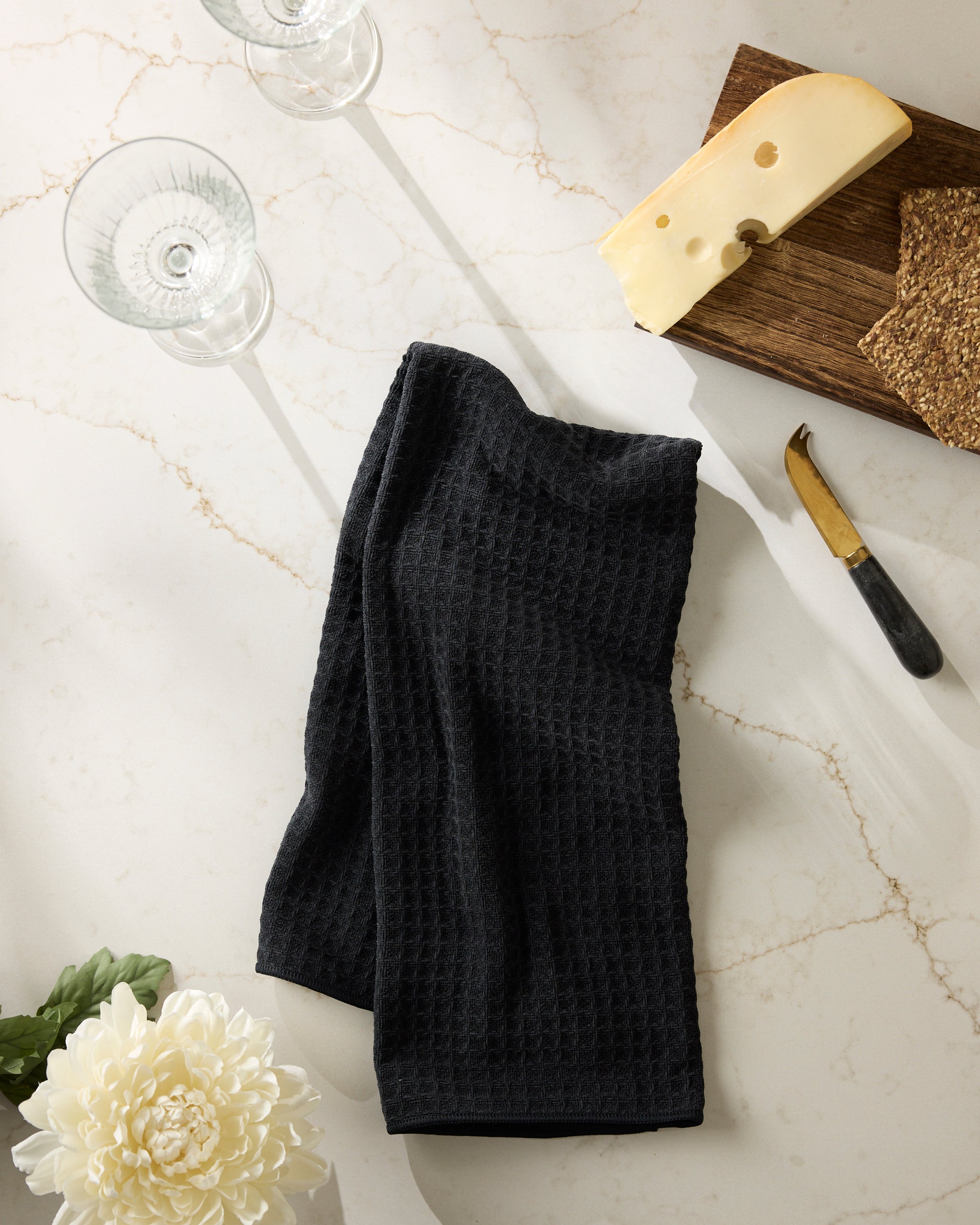 A folded Onyx black textured kitchen towel by Geometry on a marble countertop, with two empty wine glasses, a wooden board with cheese and crackers, a cheese knife, and a white chrysanthemum nearby.