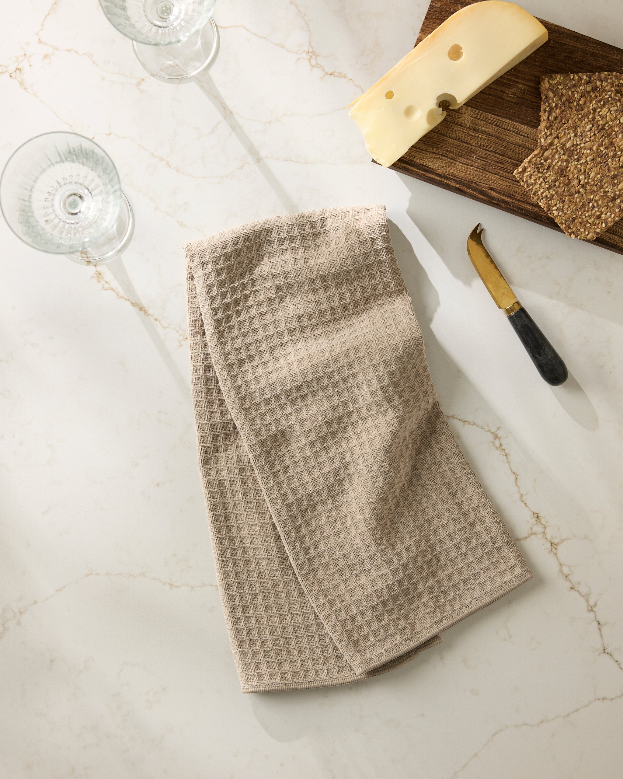 The Geometry Latte kitchen towel in beige rests on a light marble counter beside two wine glasses, a wooden cheese board with assorted cheeses, seeded crackers, and a small cheese knife.