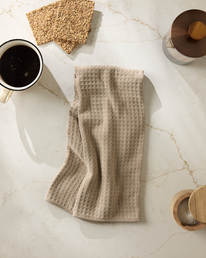 The Geometry "Latte" kitchen towel with a beige waffle texture is displayed on white marble near a cup of black coffee, crispbread, and wooden utensils in soft natural light.
