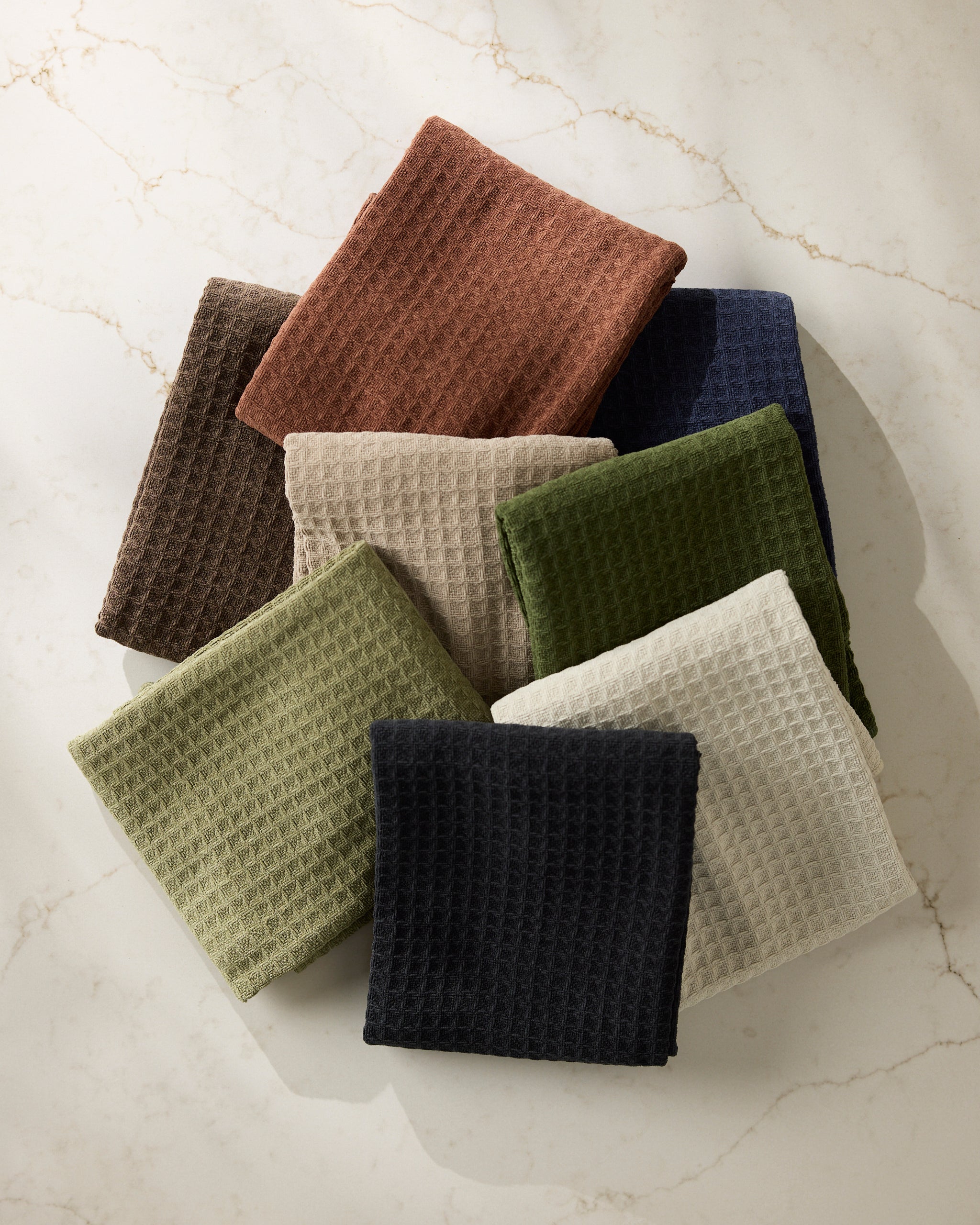 Eight folded Geometry Olive towels in brown, rust, navy, olive green, beige, cream, black, and light green are neatly arranged on a light marble surface.