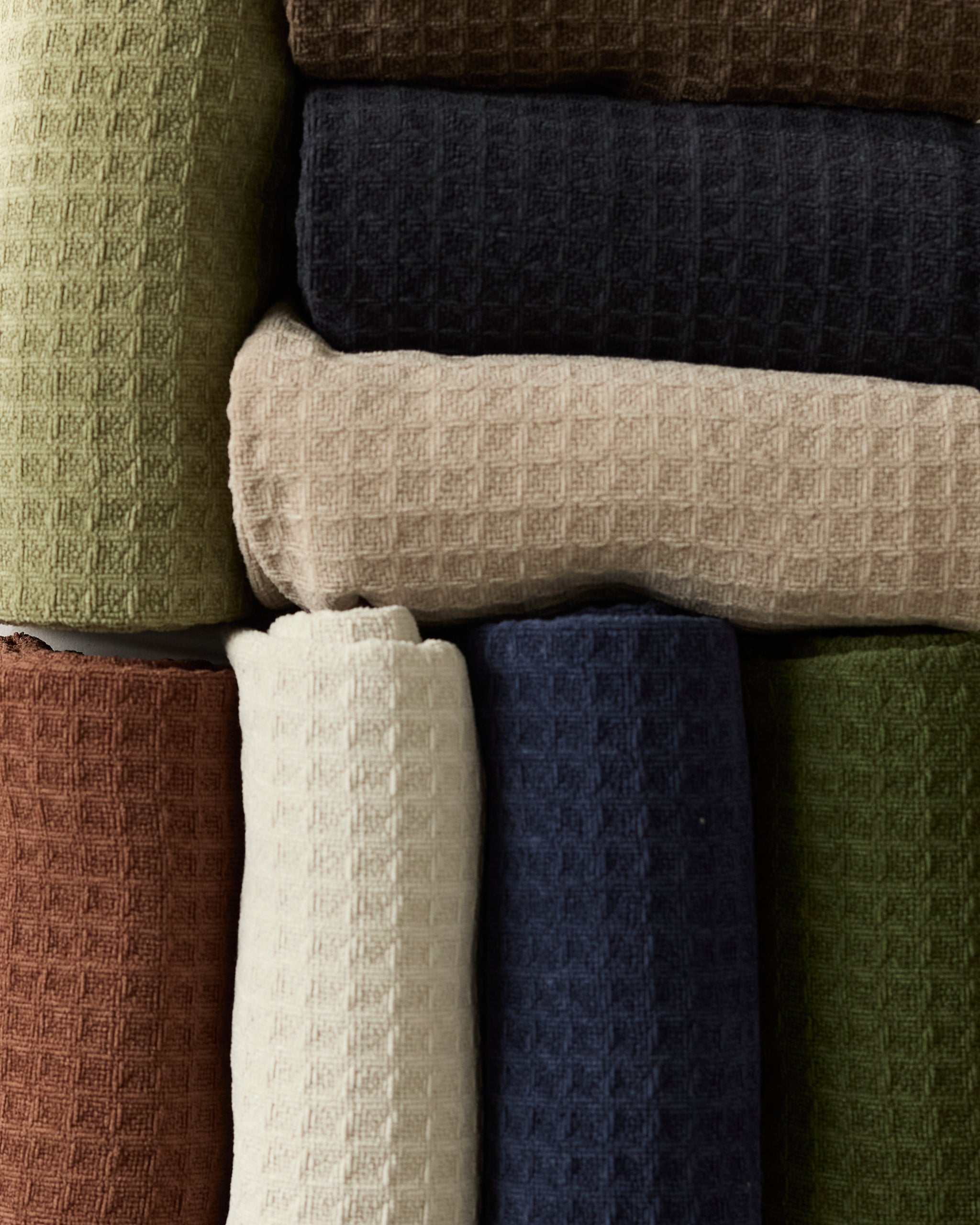 Neatly stacked rolls of "Vanilla" waffle-textured fabric by Geometry in an array of colors—green, brown, navy blue, beige, white, and rust—are arranged in two rows.