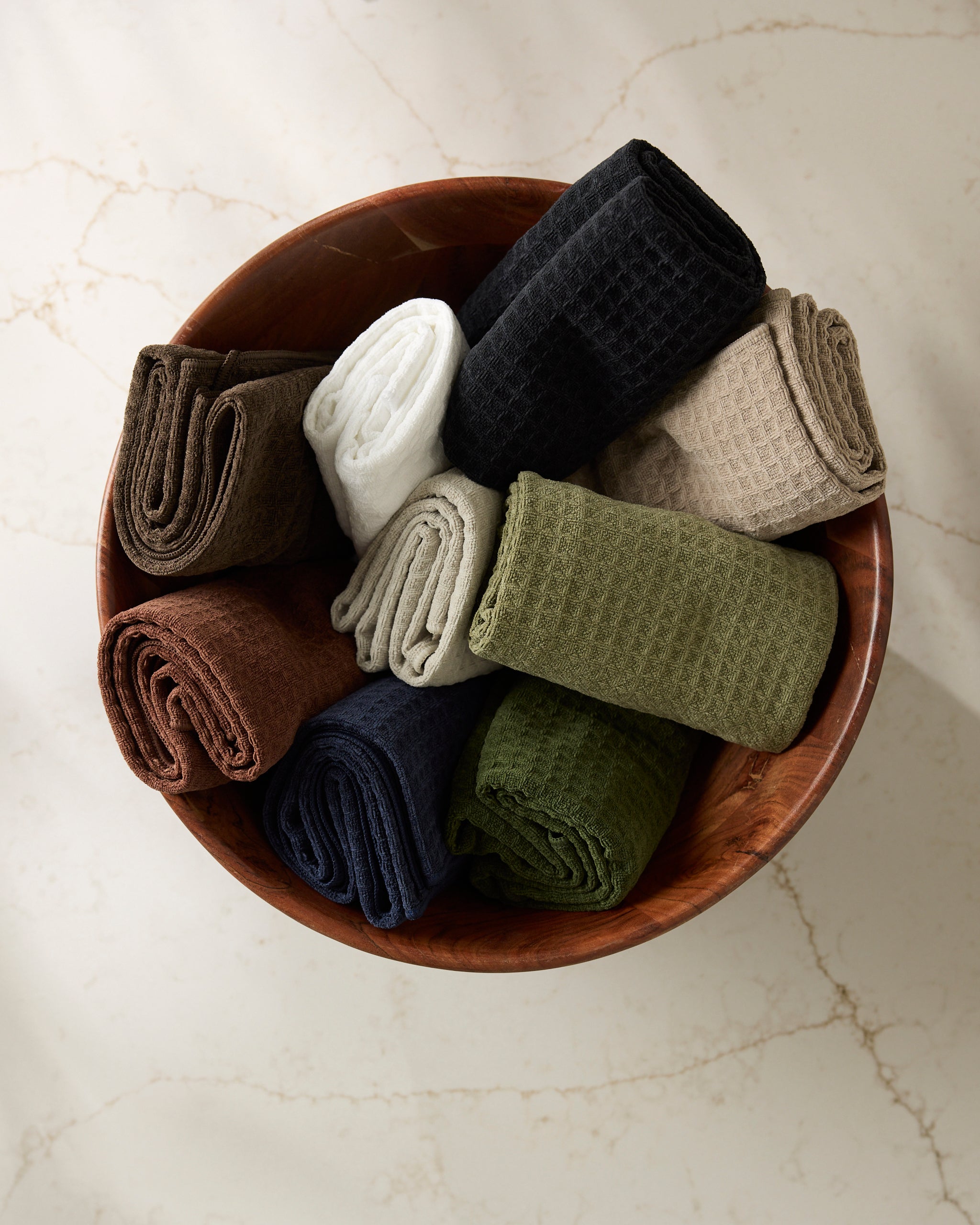 A wooden bowl holds nine Geometry Salt waffle-textured washcloths in black, white, green, navy, brown, and taupe, displayed on a light marble surface.