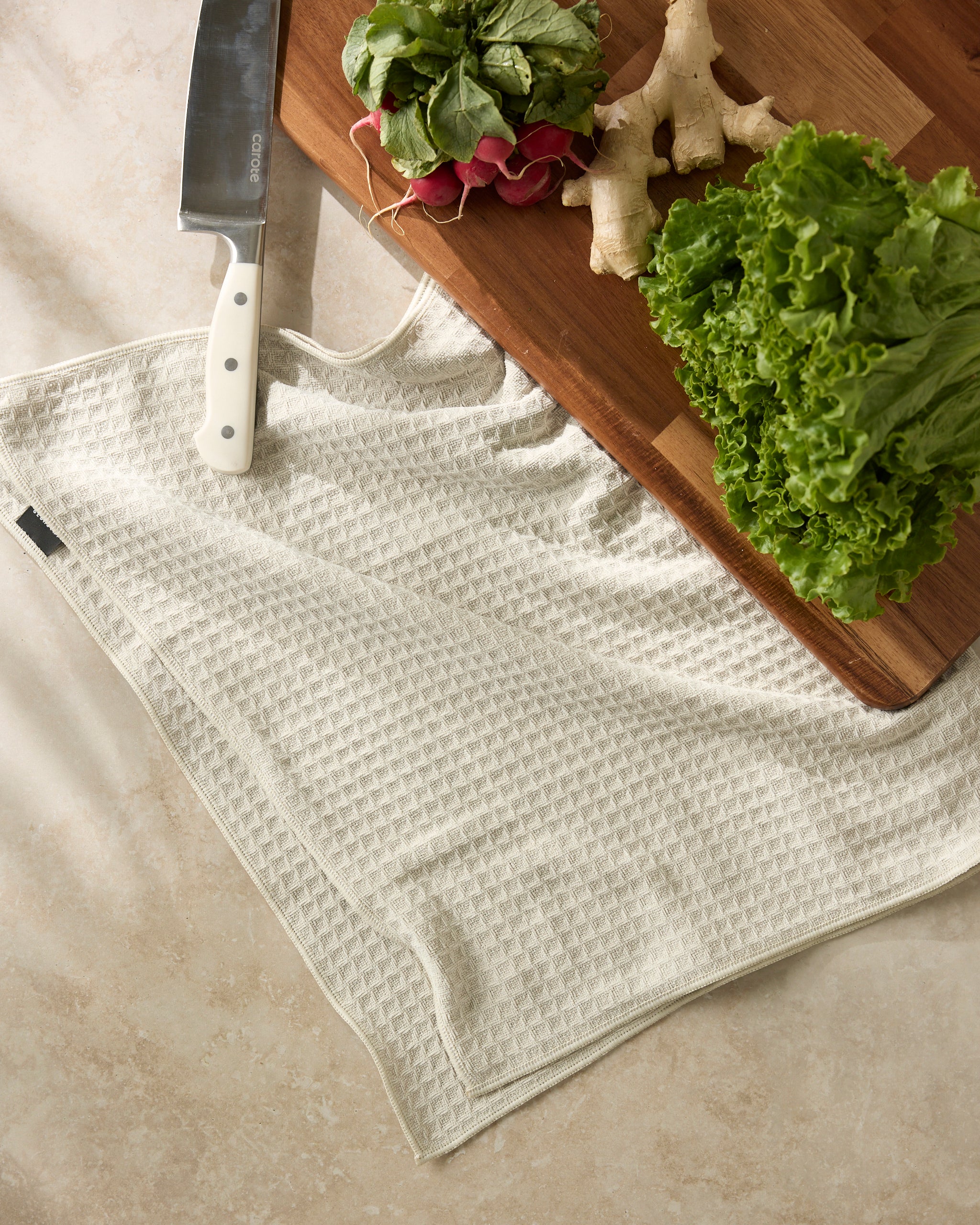 A Geometry Vanilla cutting board with leafy greens, radishes, and ginger is beside a chef’s knife on a beige textured towel on a light countertop.