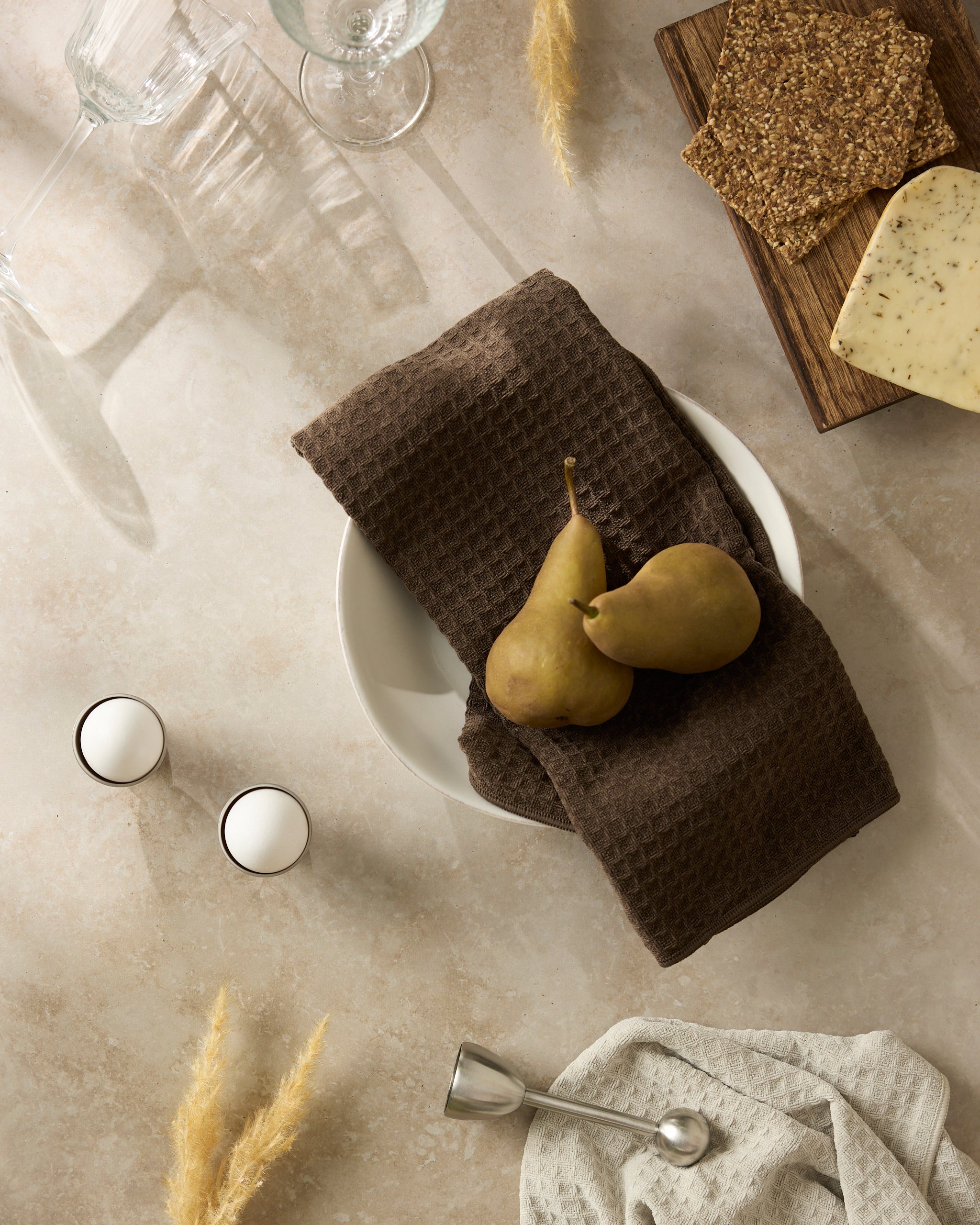 A tabletop scene featuring Geometry’s Espresso napkin displaying two pears on a white plate, beside crackers, cheese, eggs, a metal spoon, and empty glasses arranged on a light stone surface.
