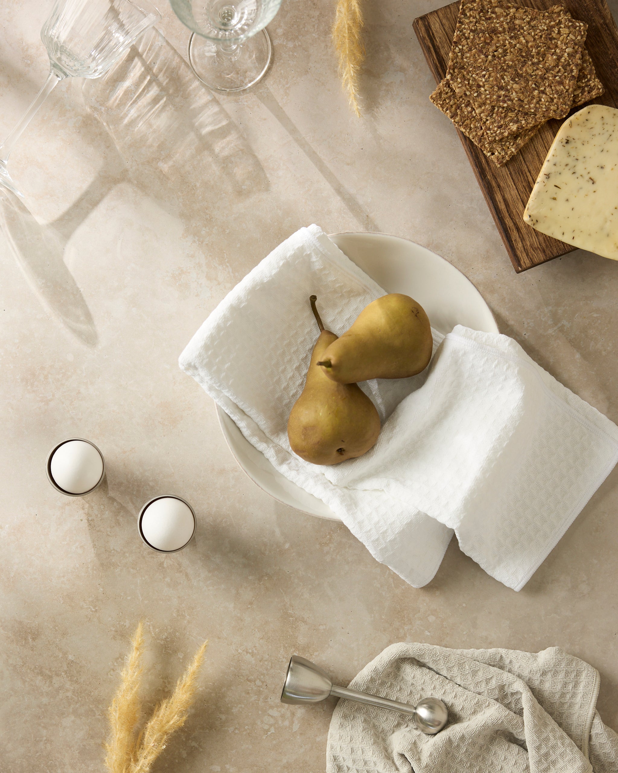 A tabletop showcases Geometry’s Salt towels, two pears on a white plate, tealight candles, cheese and crackers on a wooden board, empty glasses, dried grasses, and a metal spoon atop a textured napkin.
