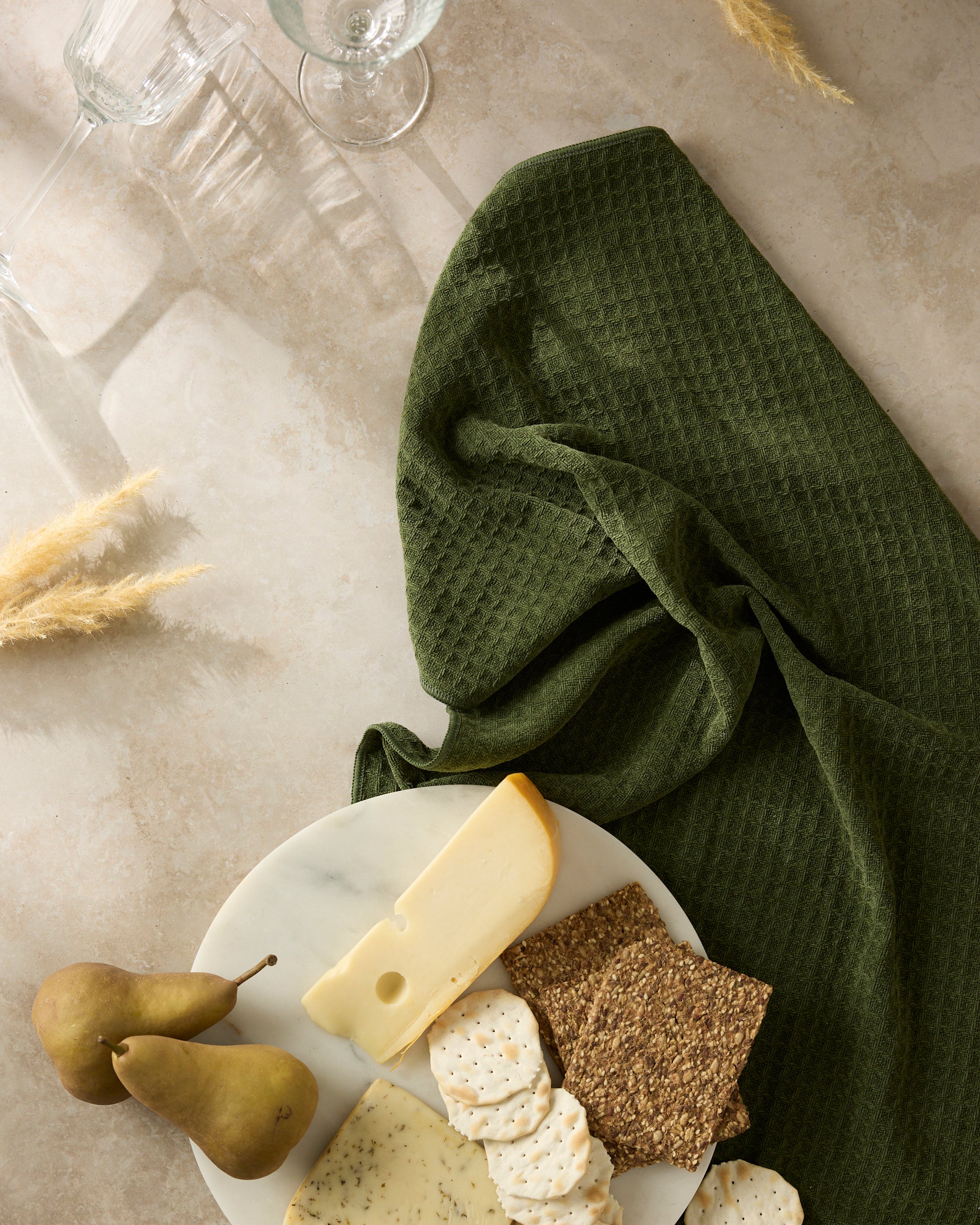 The Geometry Matcha platter displays assorted cheeses, crackers, and two pears on a beige surface beside textured dark green cloth, accented with glassware and dried grasses.