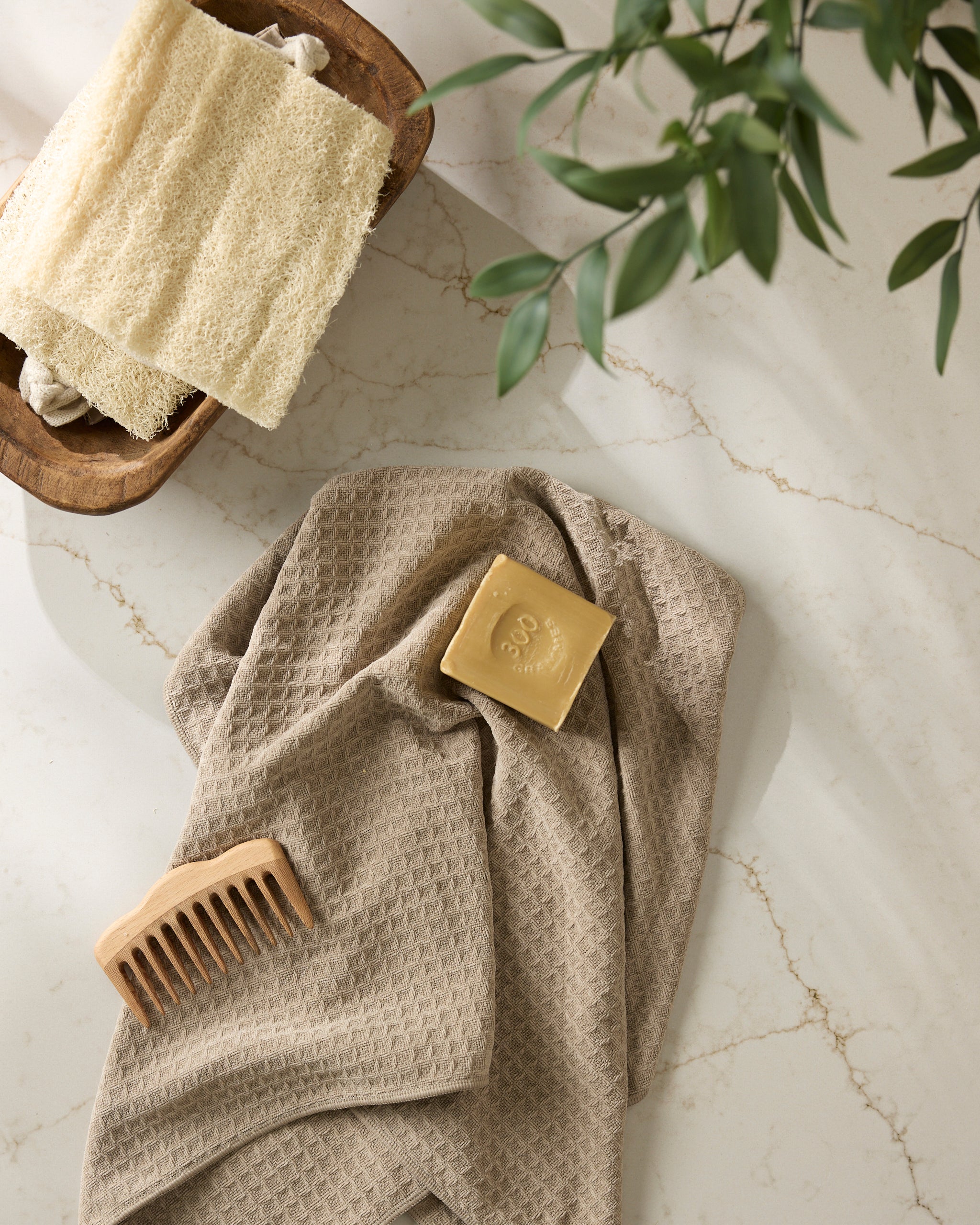 The Geometry "Latte" towel sits on marble with a bar of soap and wooden comb on top. In the corner, a wooden bowl holds a yellow washcloth and green leaves.