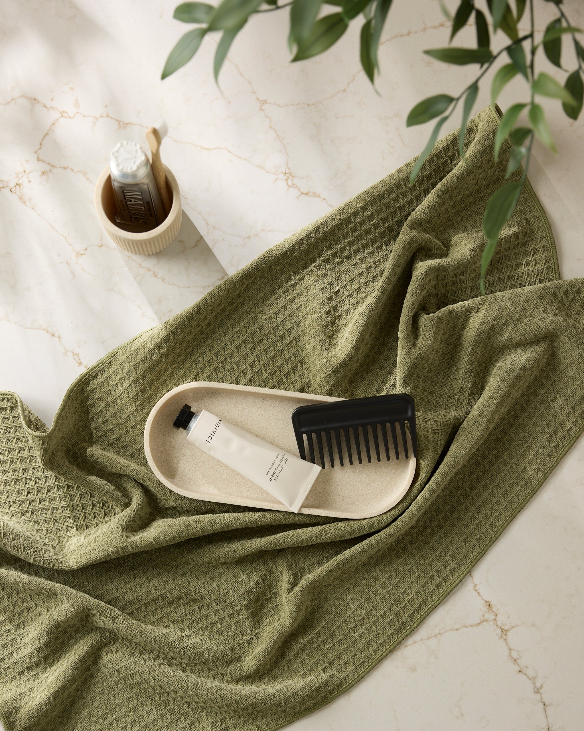 The Olive towel by Geometry, a cream tray with a black comb and white tube, and a mug holding a bottle are arranged on marble, with green plant leaves in the corner.