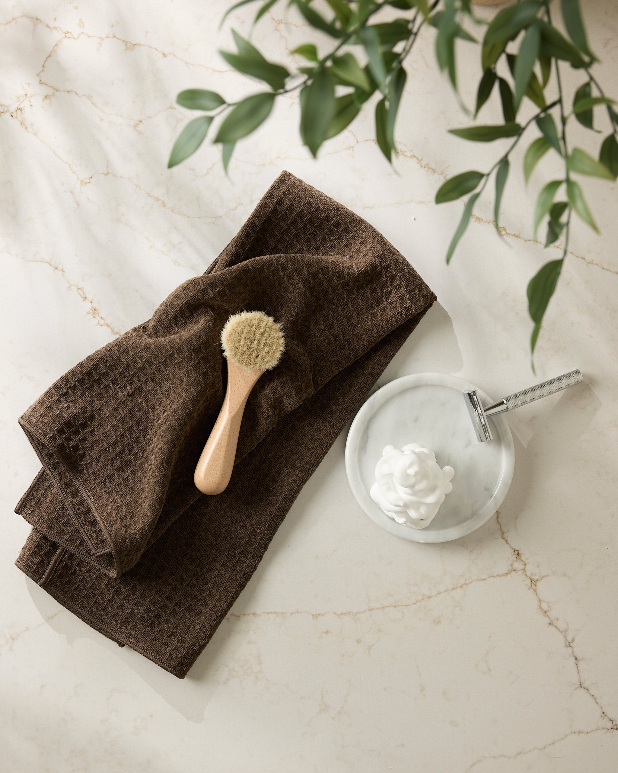 The Espresso towel by Geometry, featuring a brown textured design, is paired with a wooden bath brush, sitting on marble beside frothy soap, a silver razor, and lush green leaves overhead.