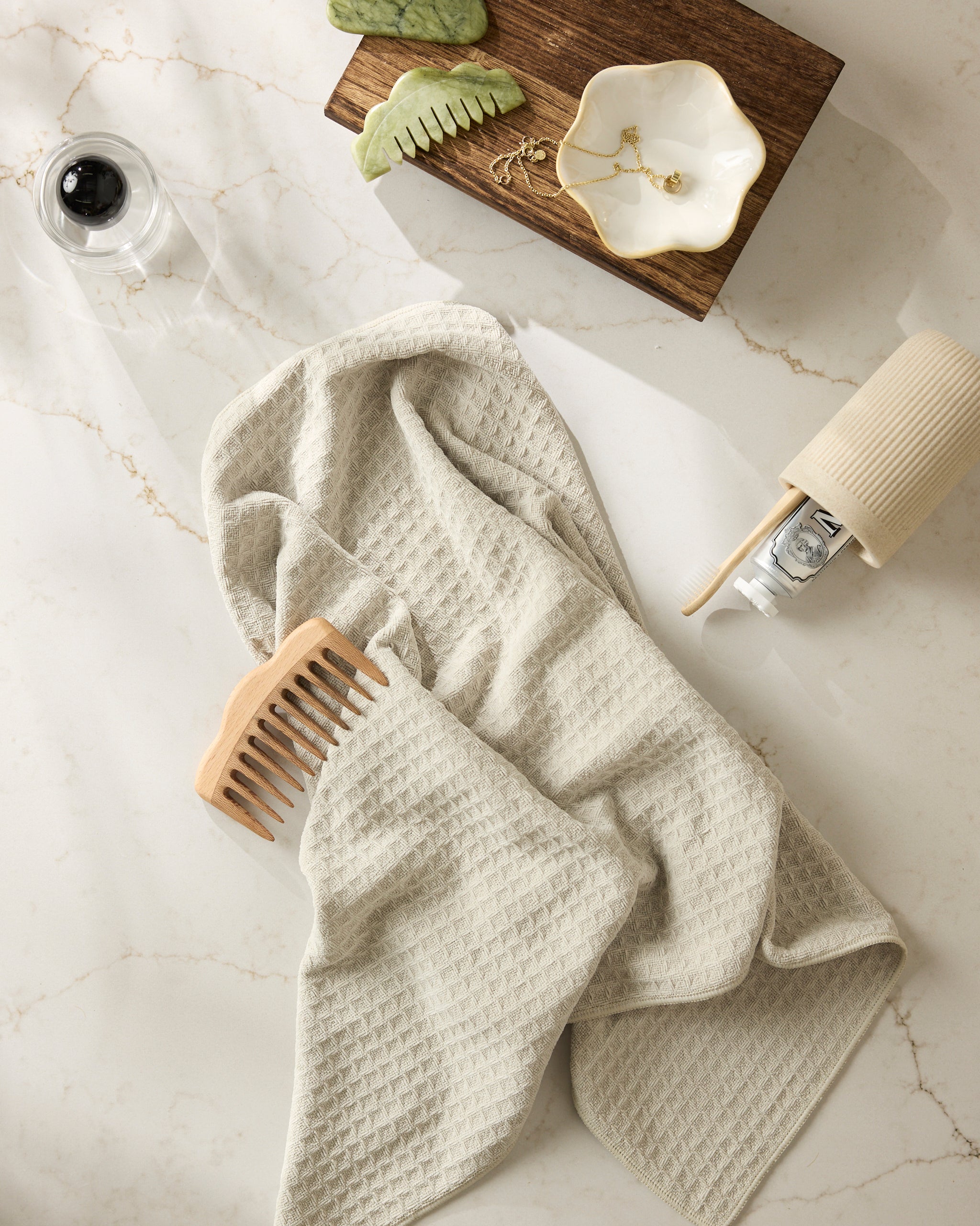The Geometry Vanilla beige waffle towel rests on marble with a wooden comb, a jade comb, jewelry in a dish, a small bottle, and a tipped cup.